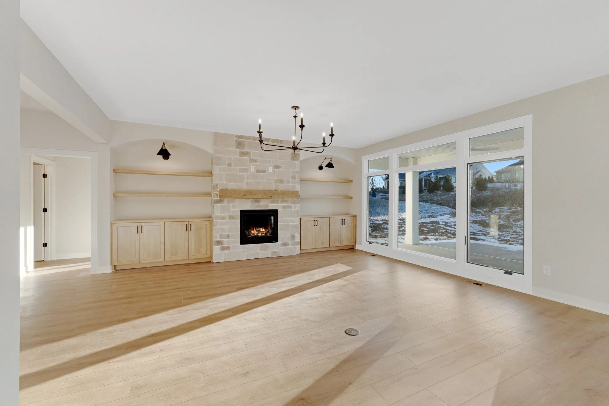 Empty living room with hardwood floors, a stone fireplace, built-in cabinets, large windows, and a chandelier.