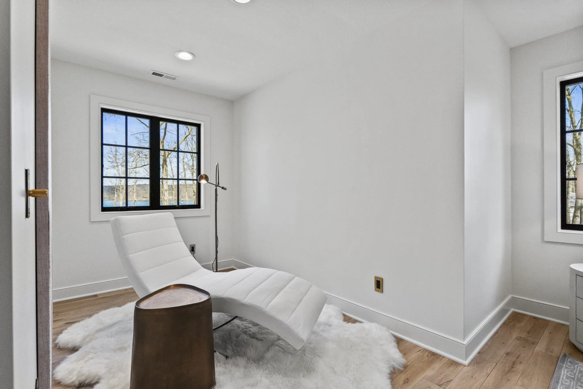 Minimalist white room with a reclining white lounge chair on a fluffy white rug, next to a small wooden side table, a tall modern floor lamp, and two black-framed windows showing a tree outside.