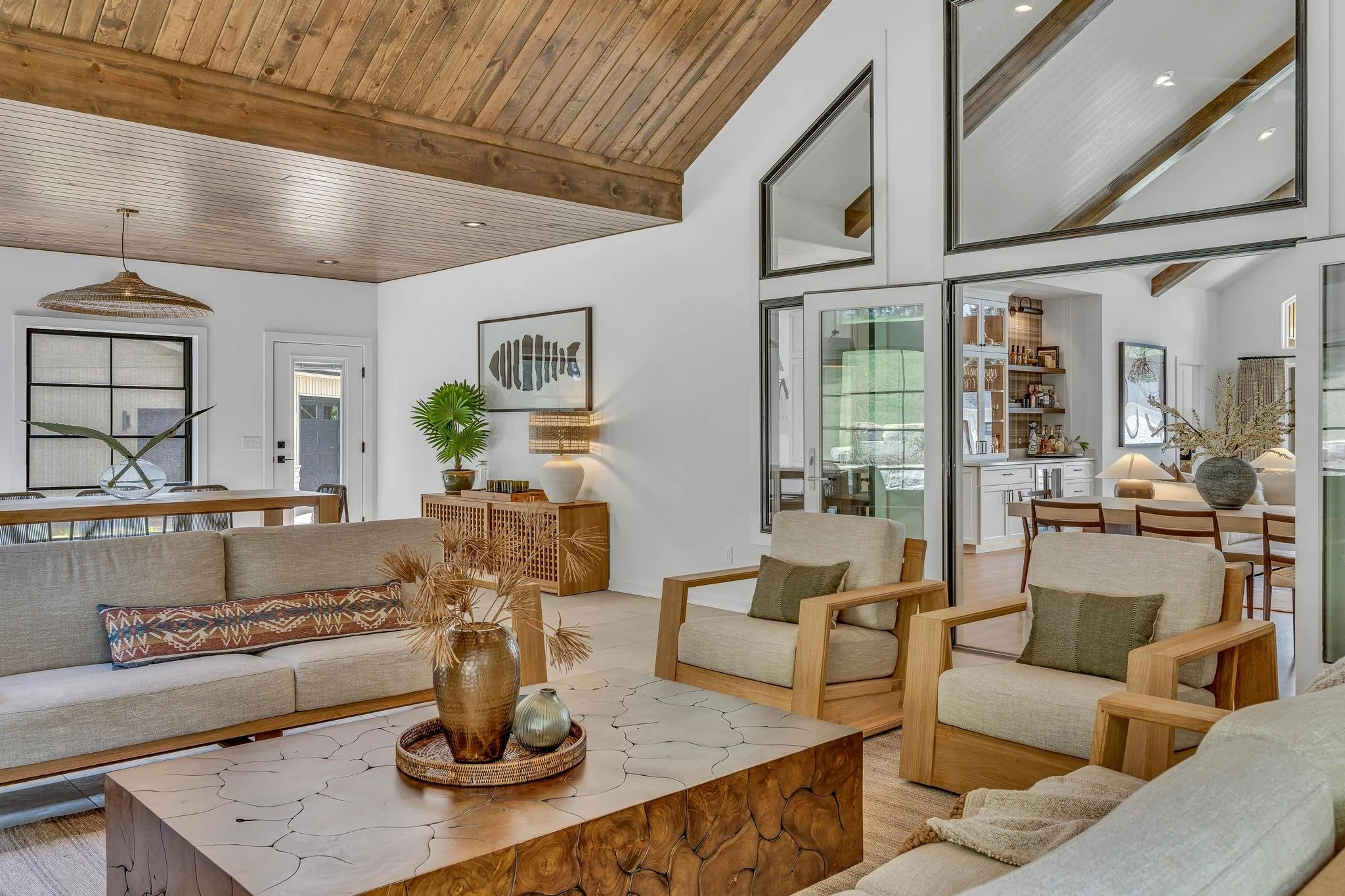 Living room with beige sofas and armchairs, wooden coffee table, decorative vases, white walls, wooden ceiling, and large windows.