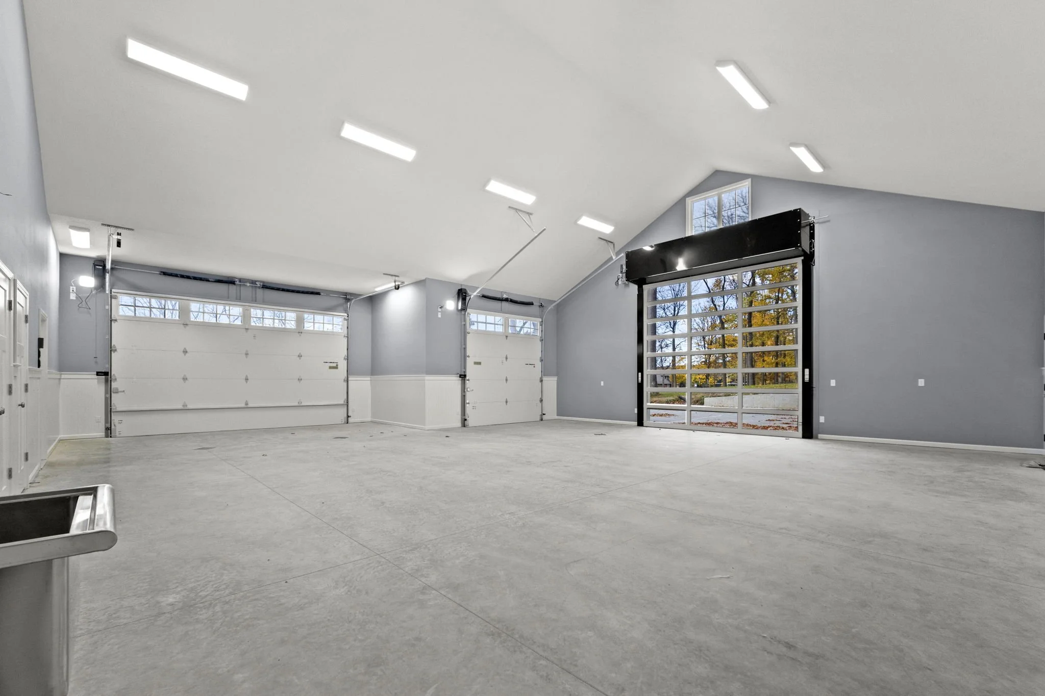 Empty spacious garage with gray walls, concrete floor, overhead lights, large garage doors, and windows showing trees outside.