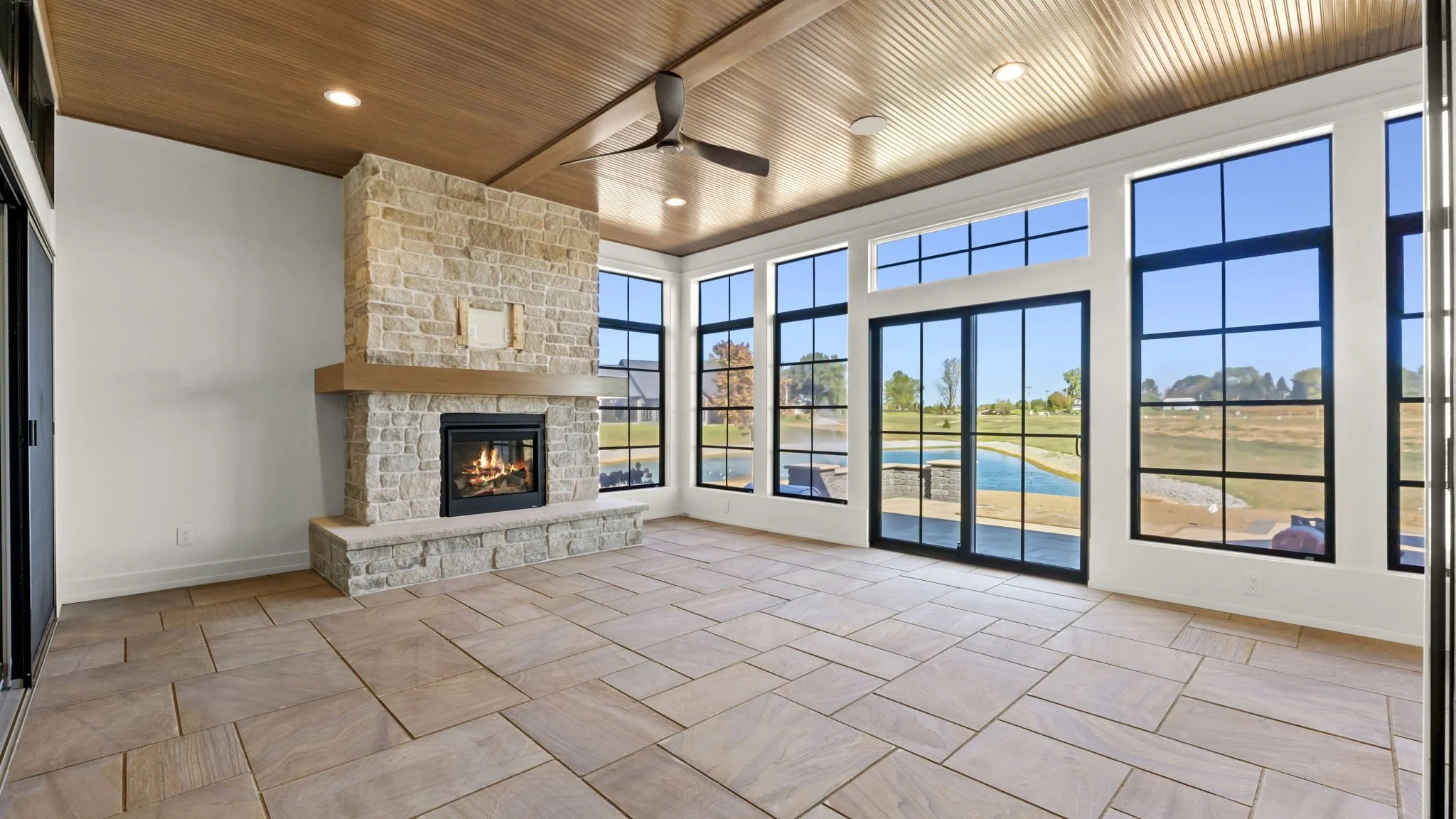 Bright sunlit indoor living room with large windows, stone fireplace, wooden ceiling with ceiling fan, and outdoor view of a pond and trees.