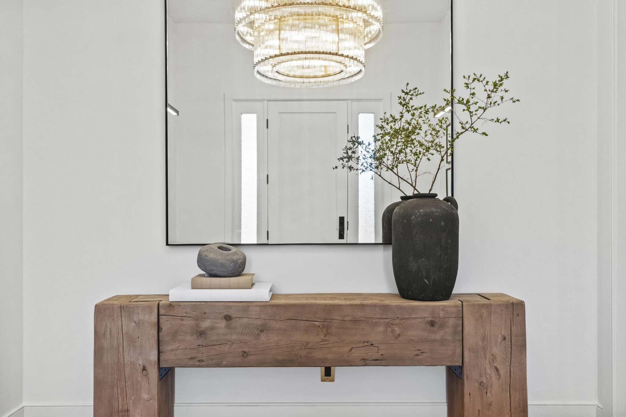 Modern entryway with a large mirror, a wooden console table, a black vase with greenery, a rock sculpture, books, and a chandelier overhead.