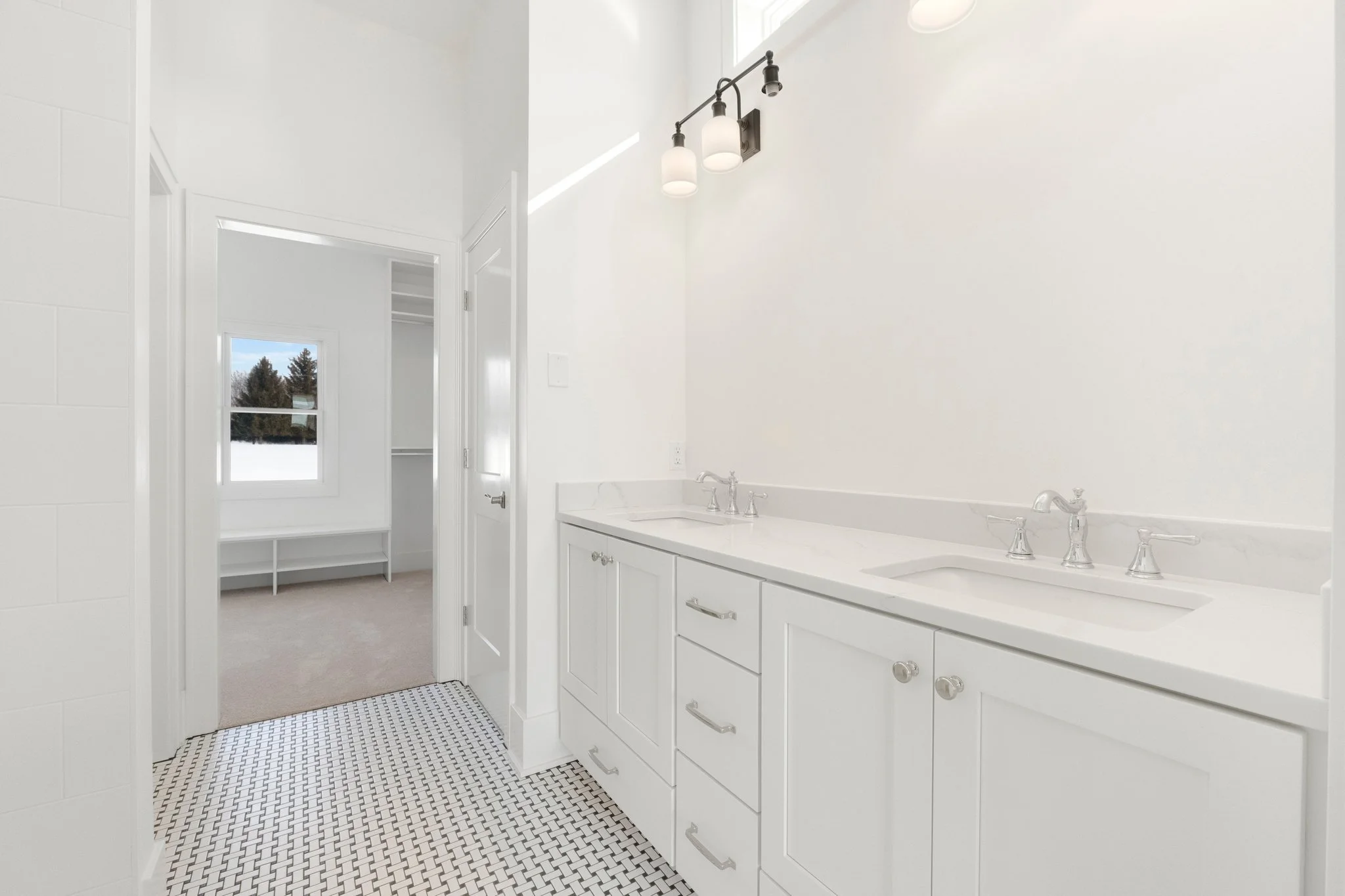Bright white bathroom with double sinks, modern fixtures, and patterned black and white tile floor. Adjacent room with window showing trees and snow outside.