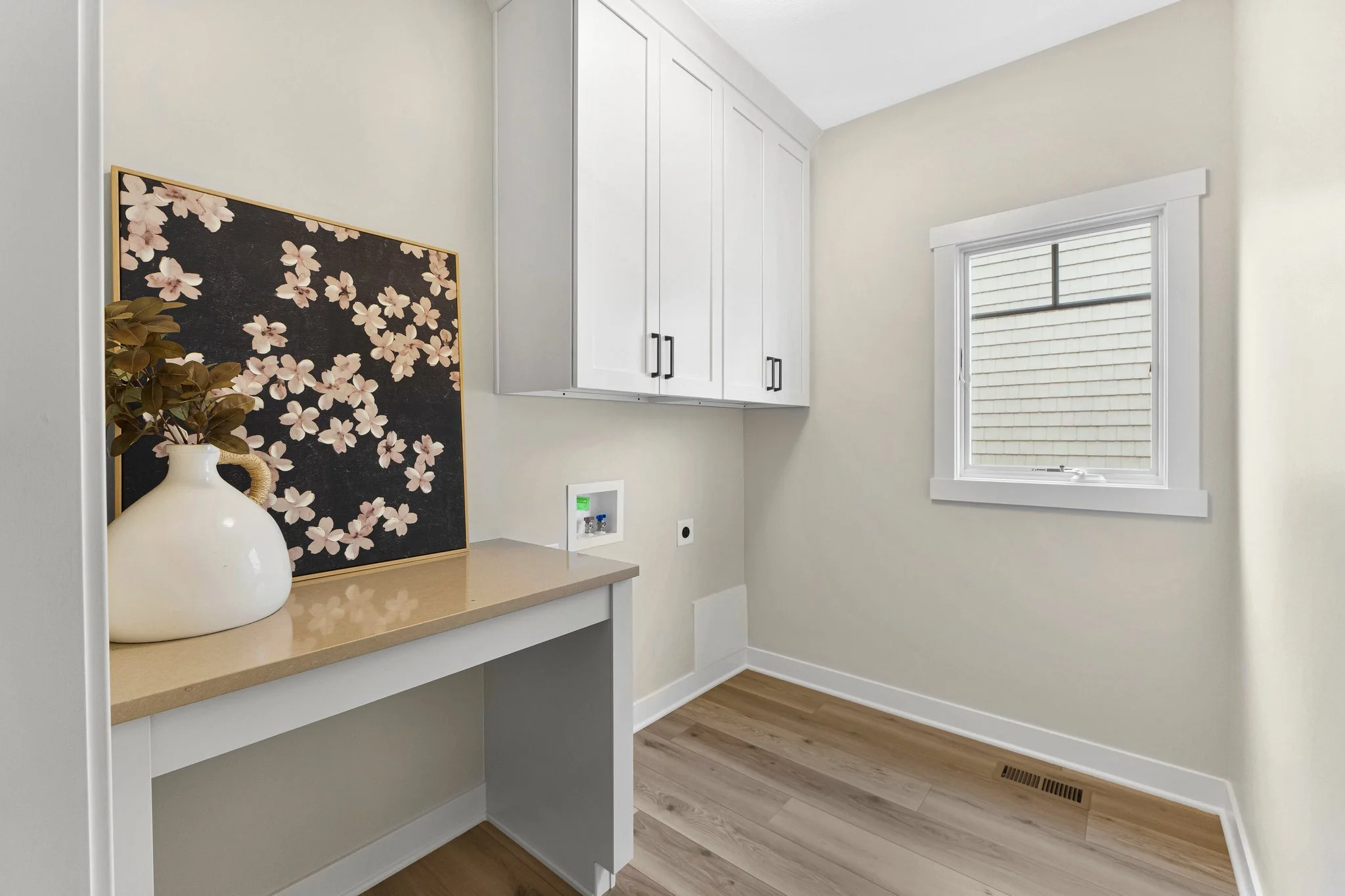 Empty laundry room with white cabinets, a window, and a beige countertop with a decorative black and pink floral artwork and a white vase with greenery.