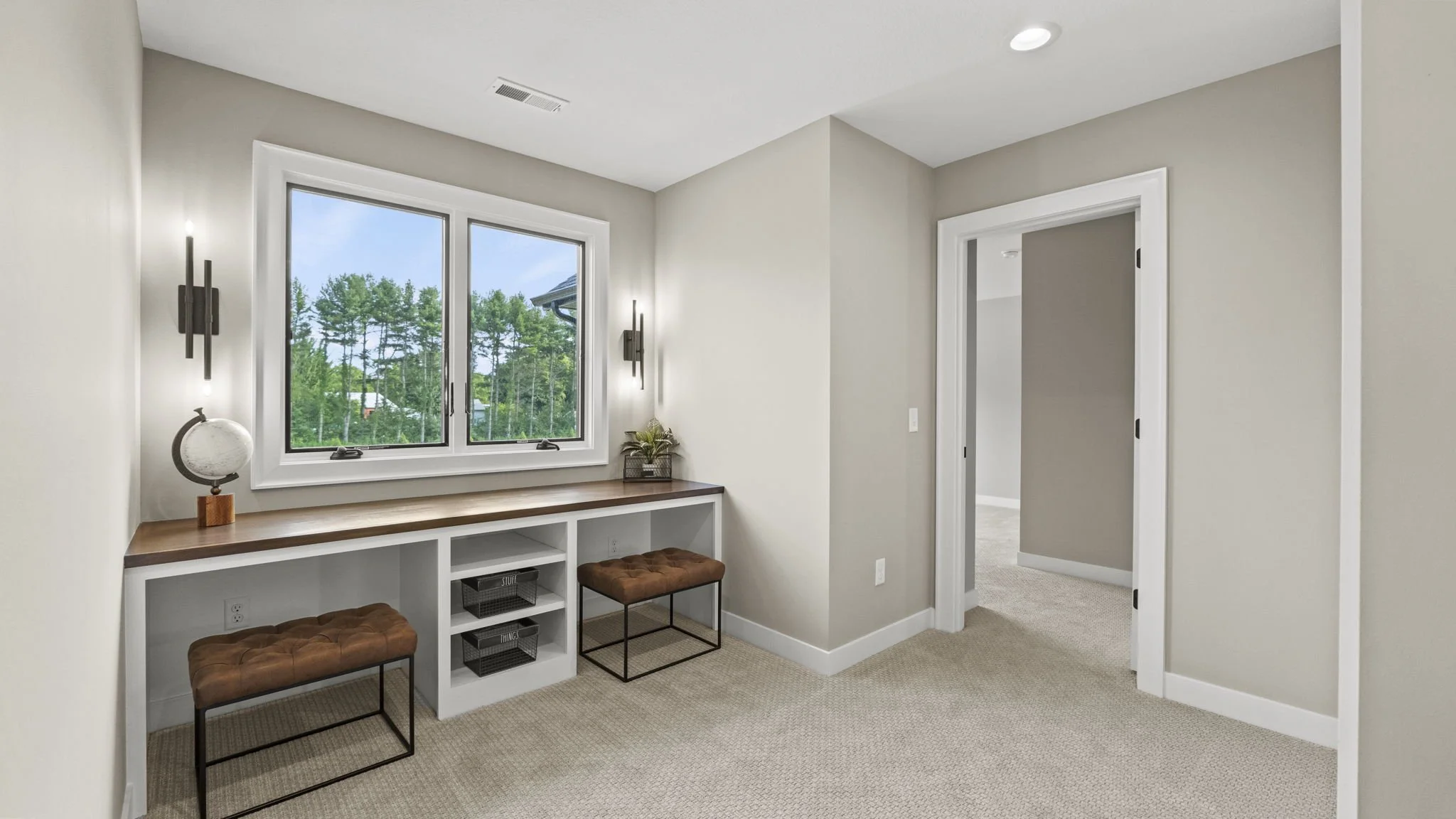 Room with built-in desk under window, two brown cushioned stools, light gray walls, beige carpet, door opening to another room, modern wall sconces, decorative plant, and globe decor.