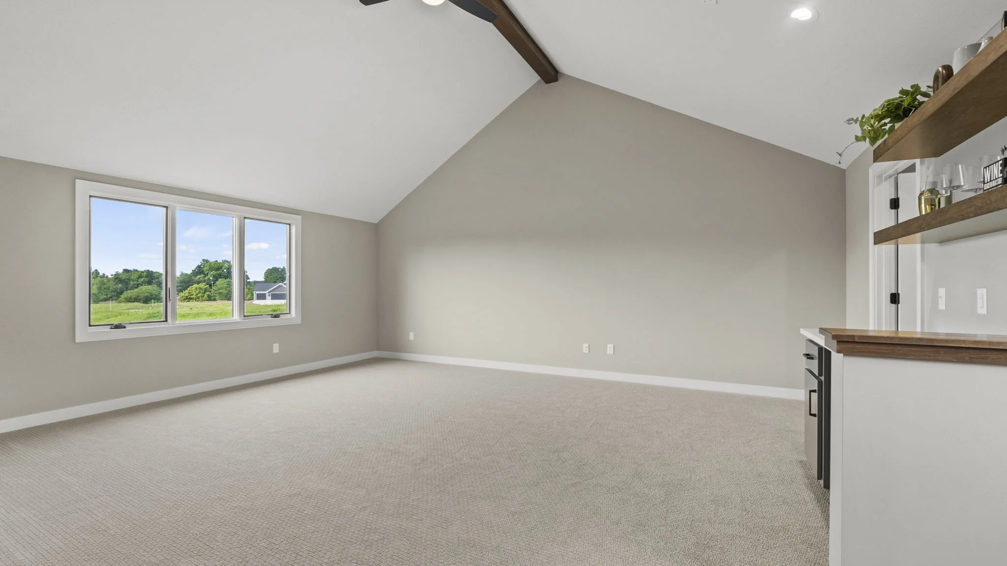 Empty room with large window, beige carpet, sloped ceiling with dark wooden beam, and partial view of a kitchen area with white and wooden shelves.