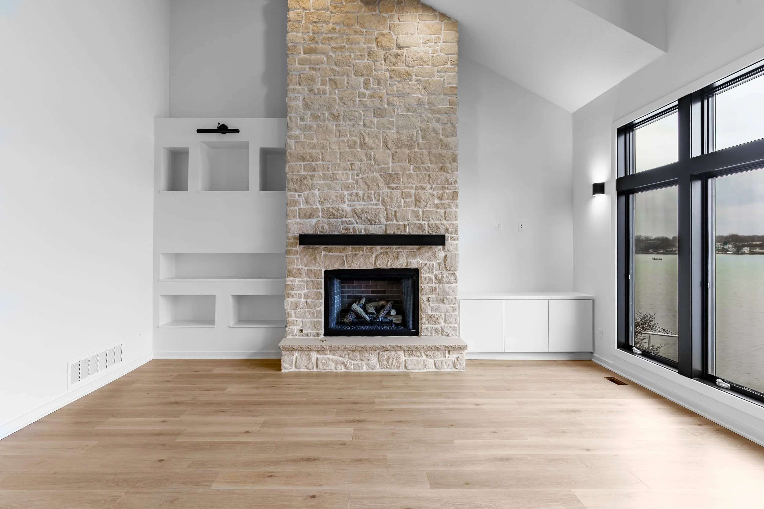 Empty living room with large windows overlooking a lake, stone fireplace, built-in white cabinets, and light hardwood floors.