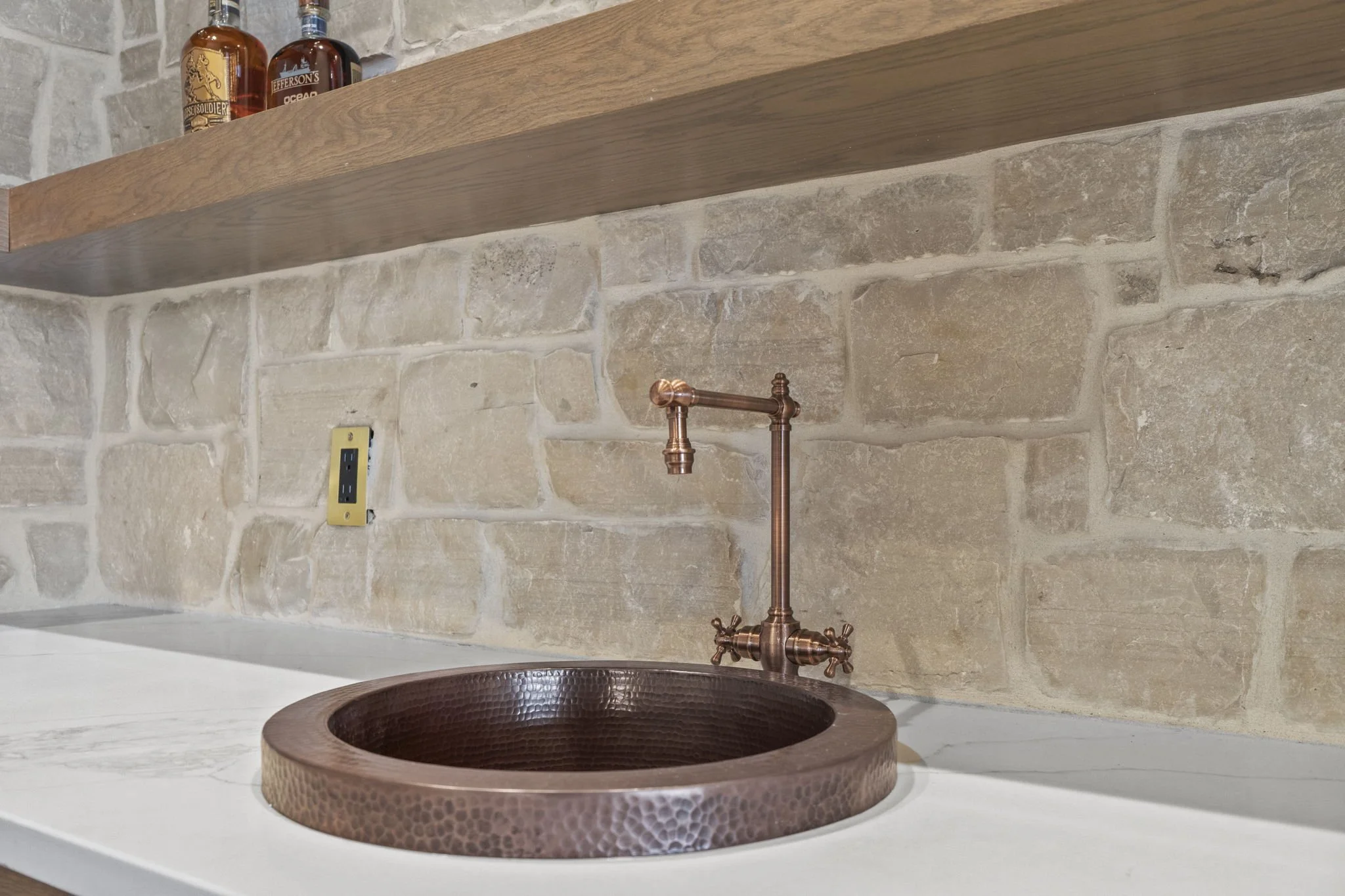 Copper hammered round sink with a copper faucet on a white countertop, beige brick wall, and a wooden shelf with liquor bottles above.