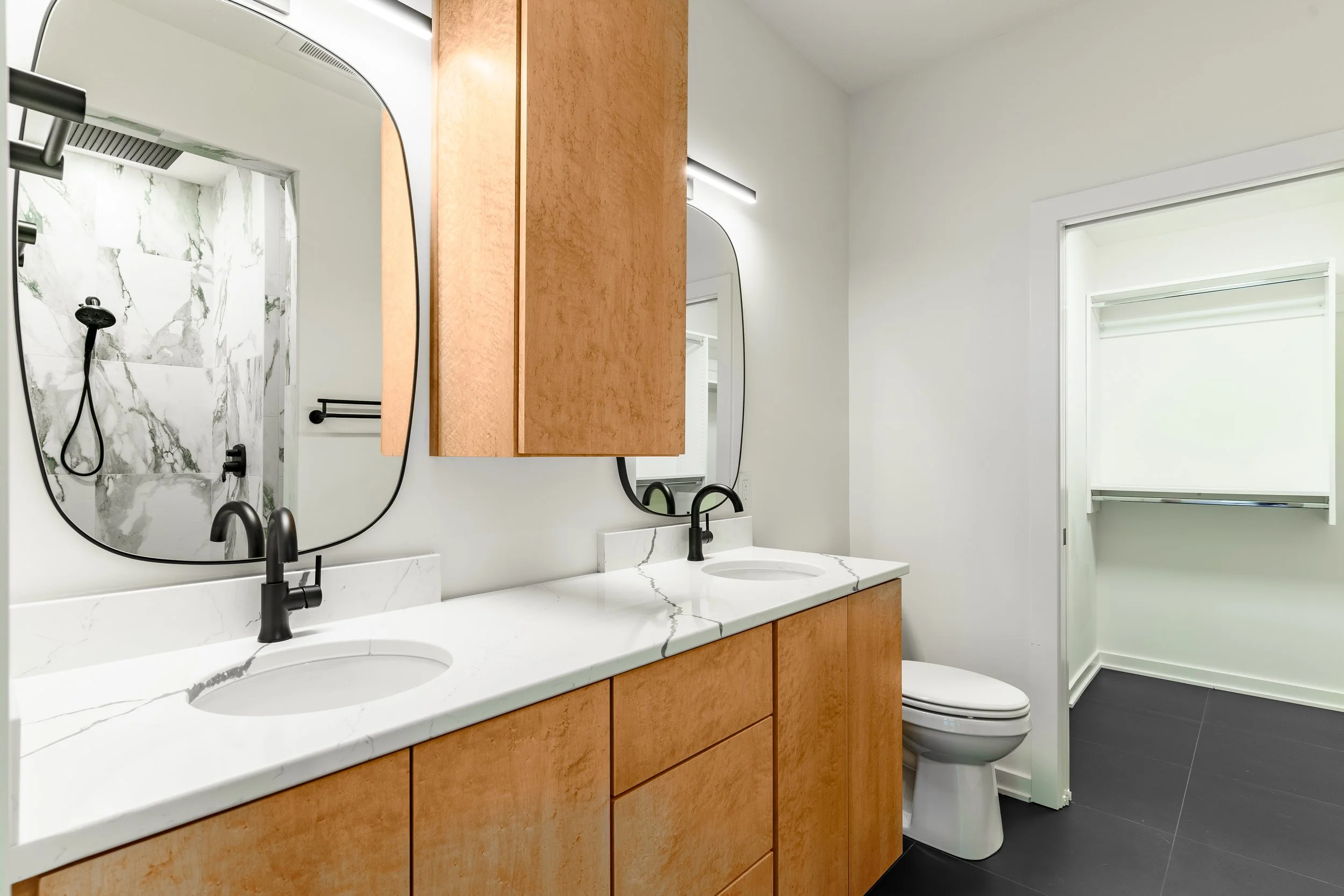 Modern bathroom with dual sinks, wooden cabinets, marble countertops, black fixtures, mirror, toilet, and a walk-in closet with a window.