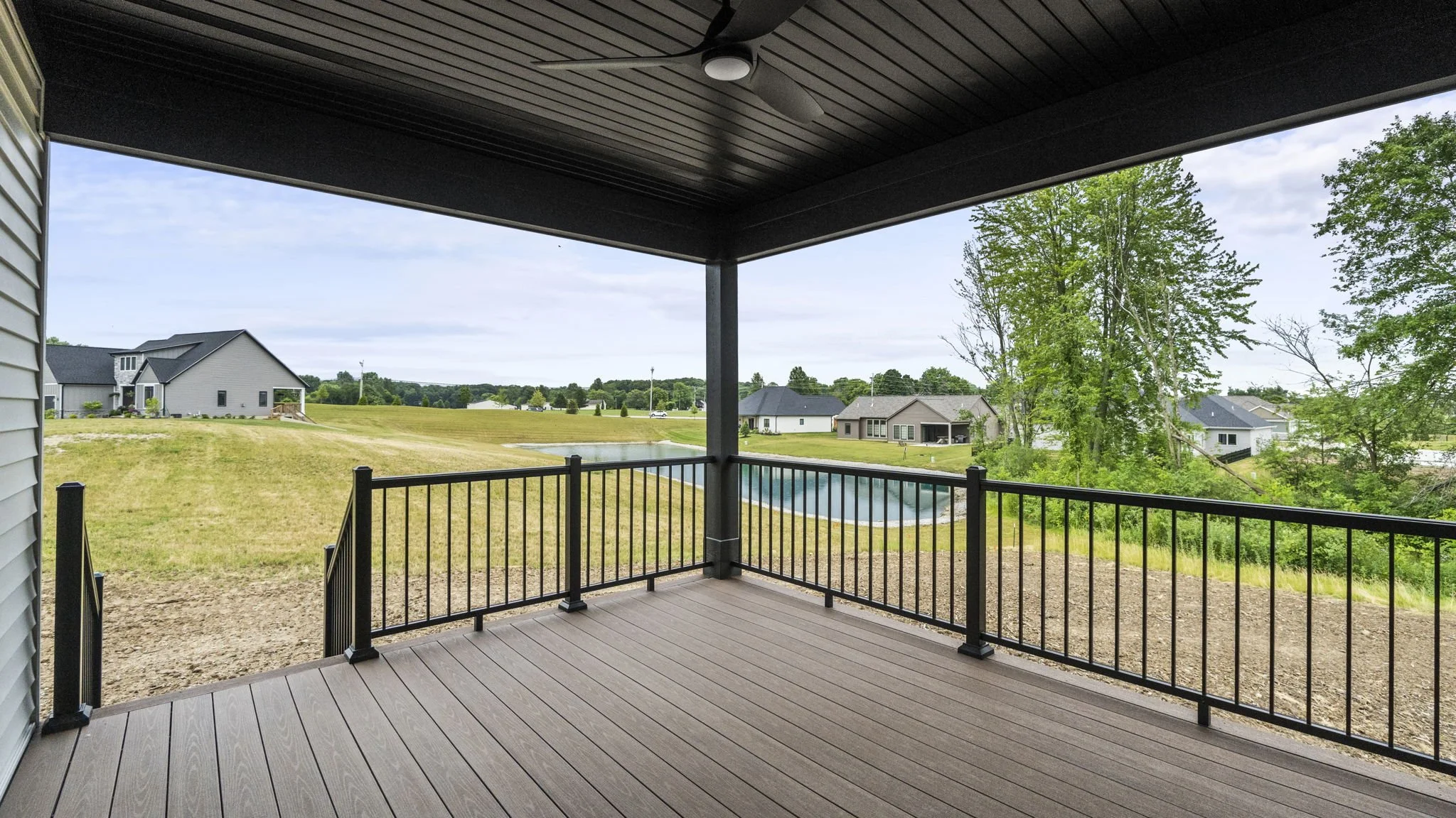 View from a covered balcony showing a grassy backyard, a pond, and neighboring houses with trees and a cloudy sky in the background.