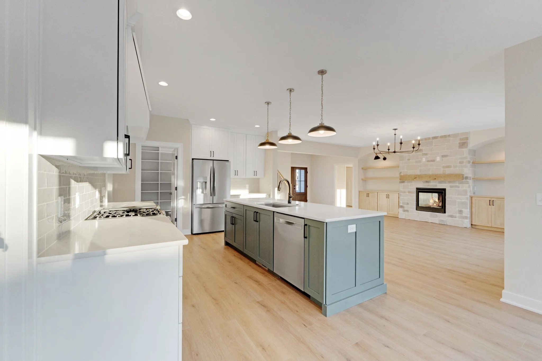 Open-concept kitchen and living room with a central island, white cabinets, stainless steel appliances, a stone fireplace, wooden flooring, and modern lighting fixtures.