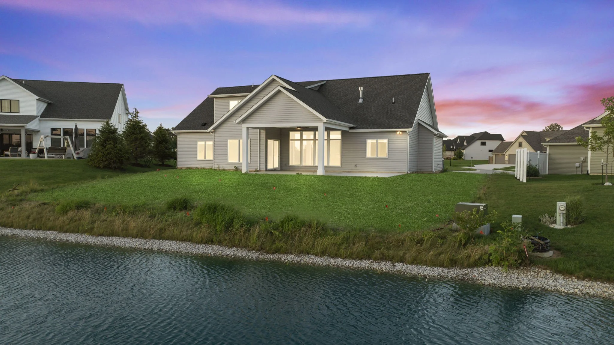 A modern house sits on a grassy lawn by a lake at sunset, with neighboring houses in the background.