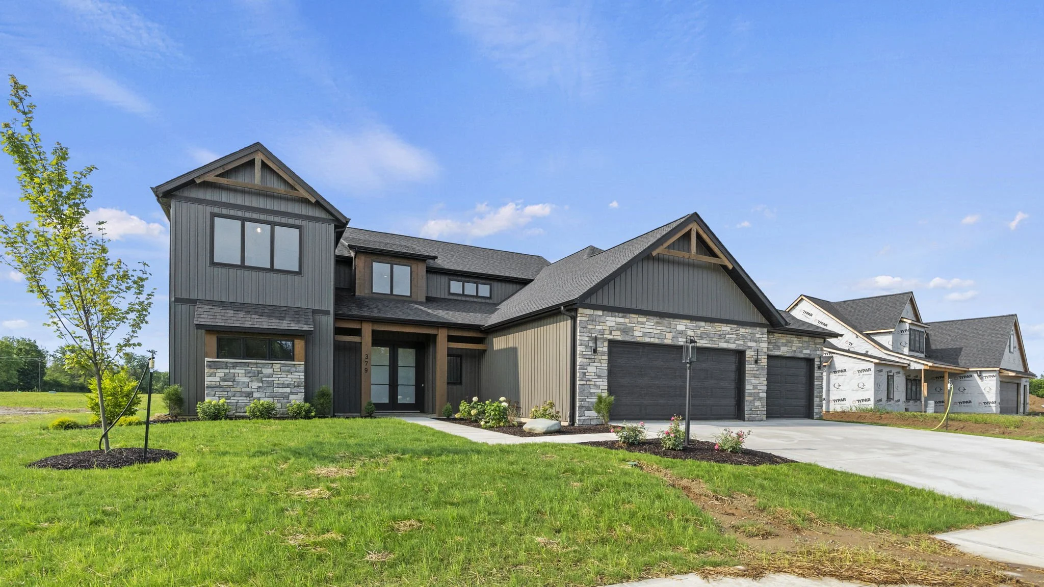 Newly built modern house with gray siding, stone accents, and a large driveway, under a blue sky with a few clouds.
