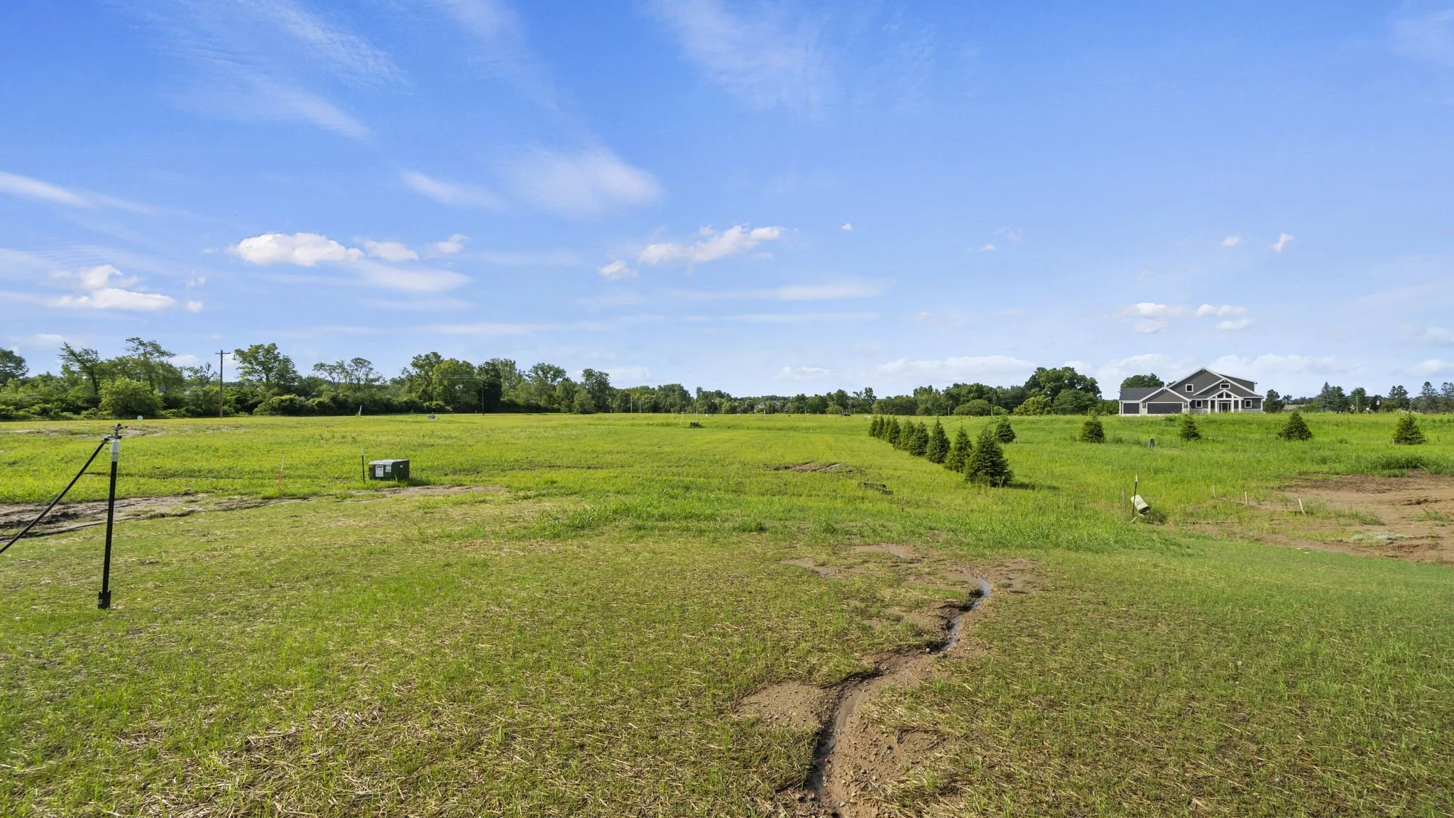 Open grassy field with small homes, blue sky with clouds, and landscape, likely in a rural or suburban area.