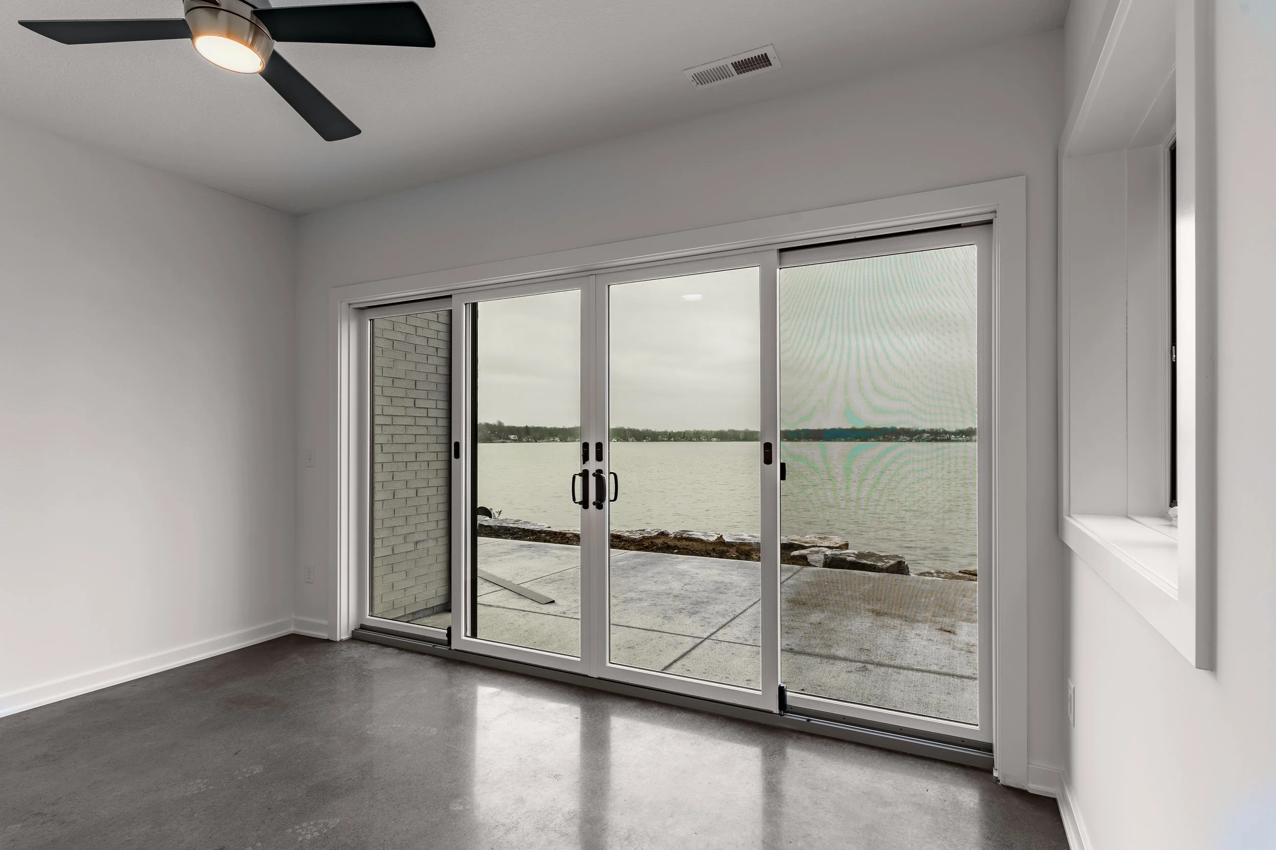 Empty room with white walls, concrete floor, ceiling fan, and large sliding glass doors opening to a lakeside view with water and cloudy sky.