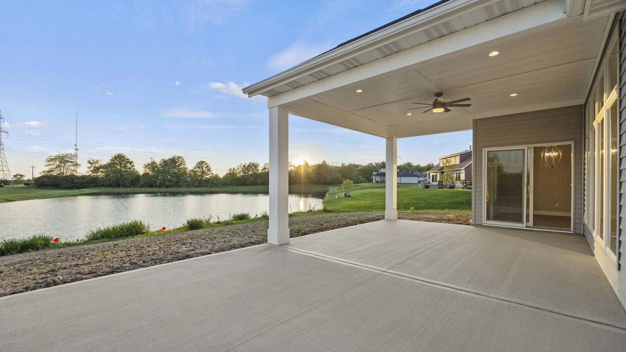 View of a backyard patio with a concrete floor and a view of a pond and green lawn at sunset. The patio has a ceiling fan and is attached to a house with sliding glass doors.