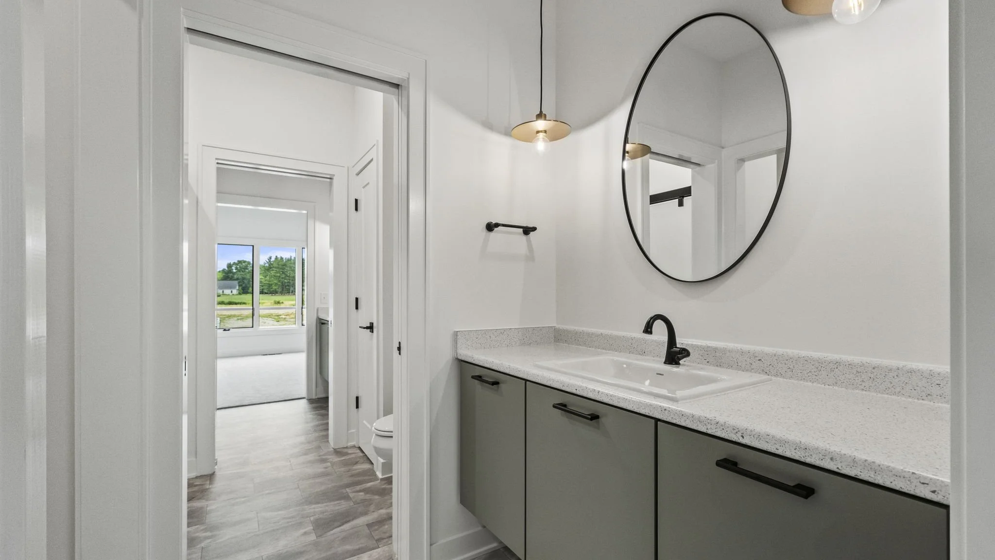 Modern bathroom with a green vanity, black faucet, round mirror, and pendant light, adjacent to a room with large windows showing a green outdoor landscape.