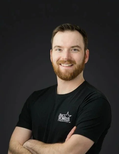 A man with short brown hair, a beard, and blue eyes smiling. He is wearing a black t-shirt with a logo on the left side that says 'Star HOMES' and some smaller text underneath, against a dark background.