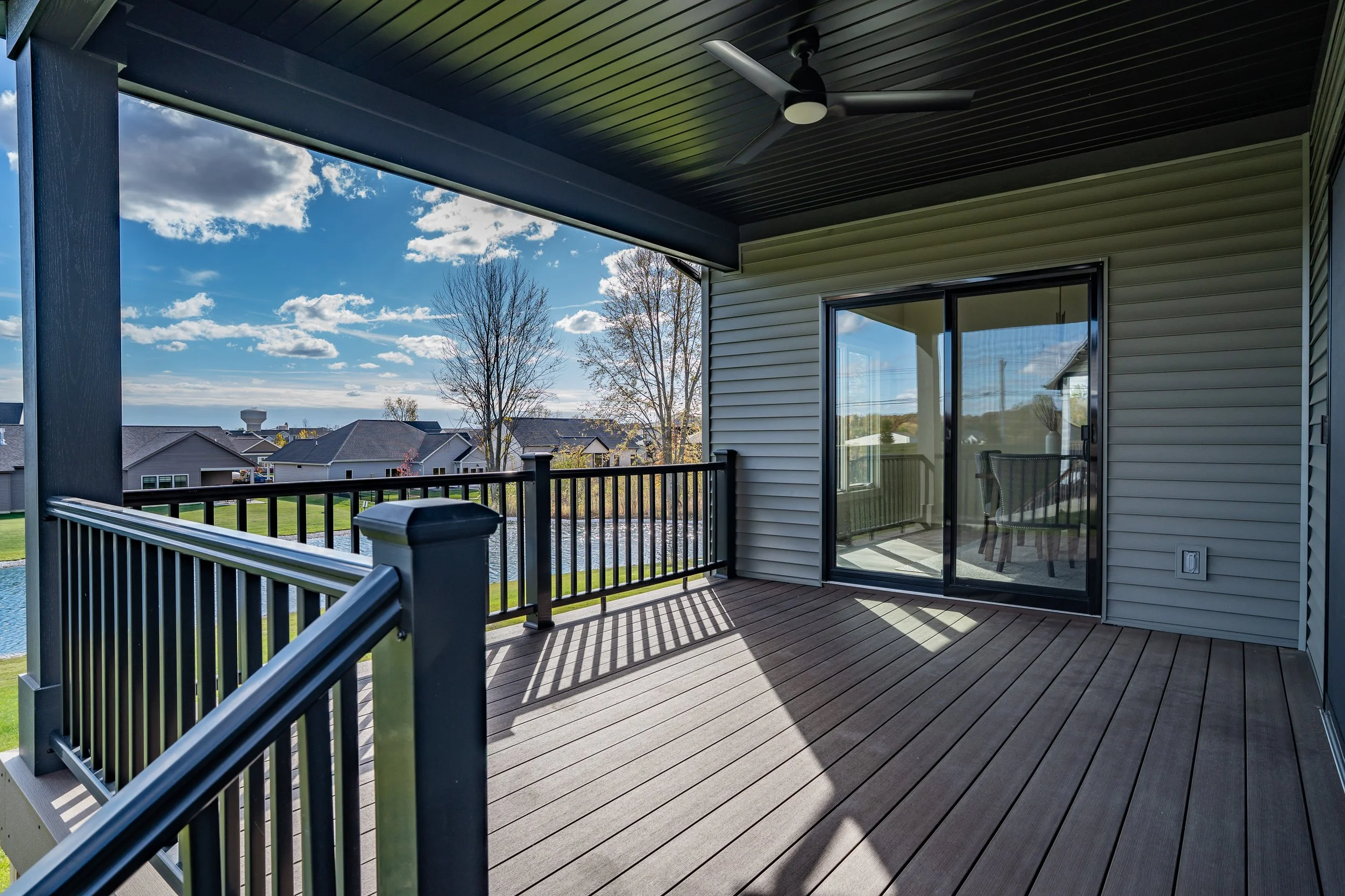 View of a balcony with wooden flooring, metal railing, and a sliding glass door leading inside. The balcony has a ceiling fan and overlooks a neighborhood with houses, trees, a pond, and a blue sky with scattered clouds.