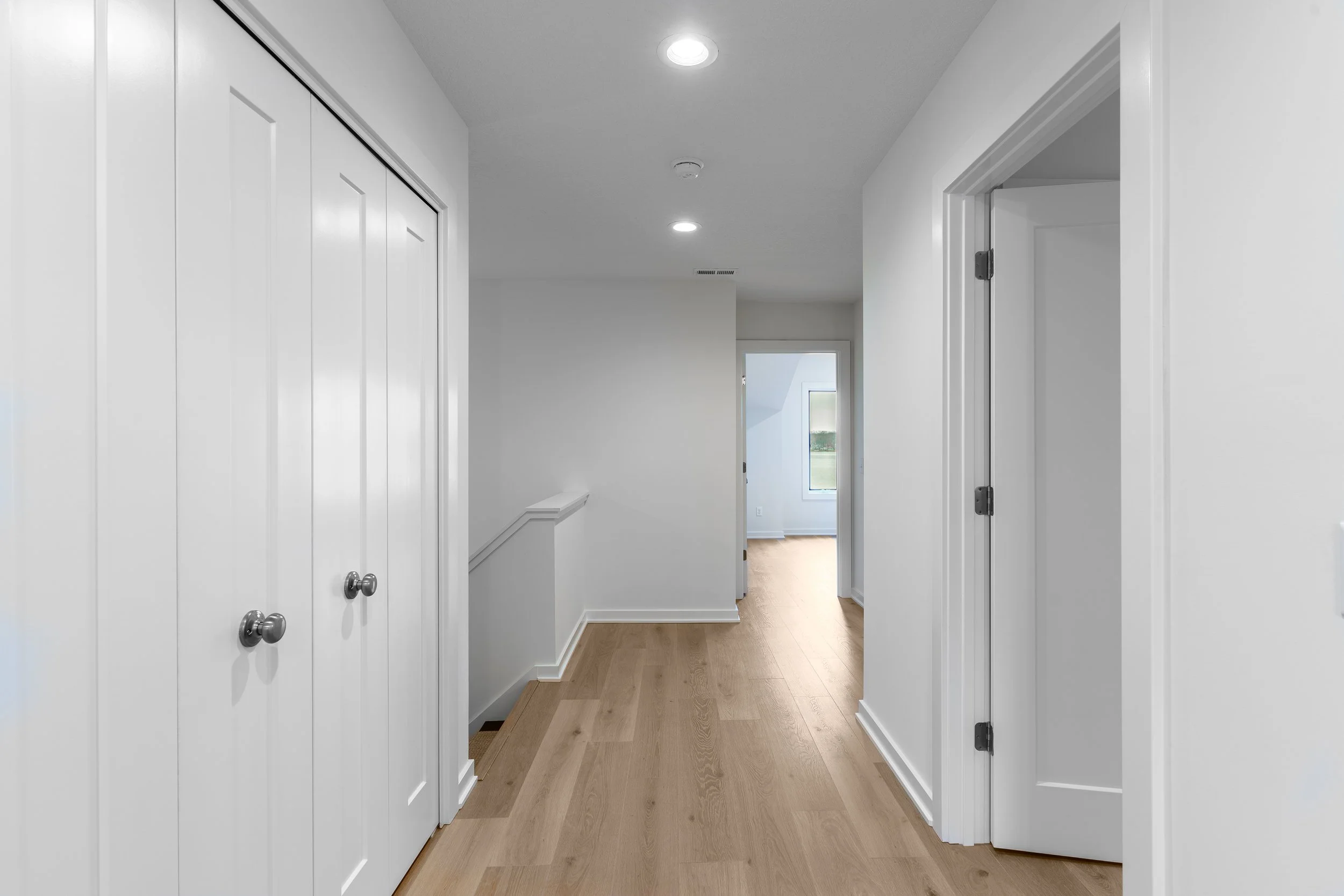 Empty hallway with white walls, wooden flooring, and ceiling lights, leading to a room with a window.