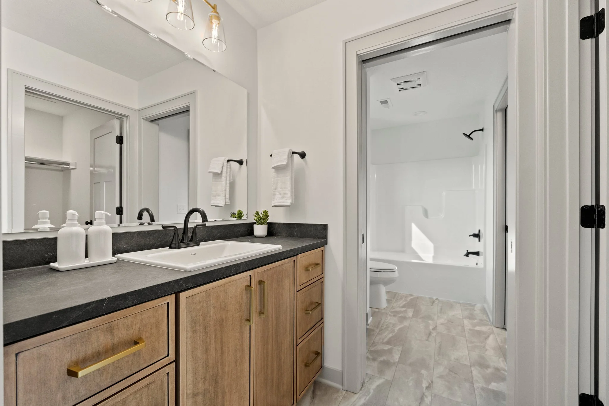 A modern bathroom with a vanity featuring a black countertop, four white soap dispensers, and a small potted plant. There is a large mirror above the vanity, a towel rack with two white towels, and a glass-enclosed shower with a black handle. The bat