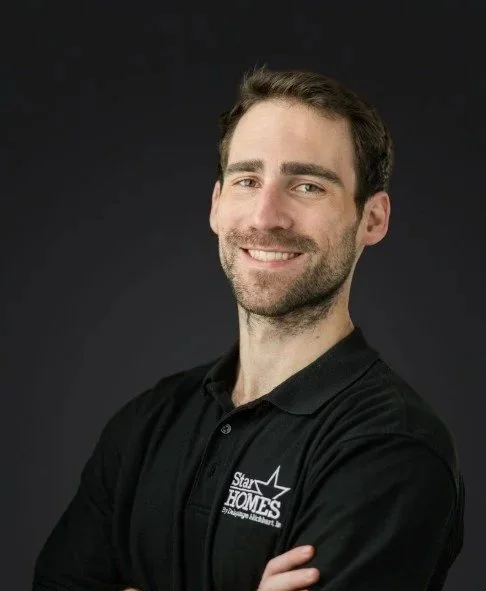 Portrait of a smiling man with brown hair and beard, wearing a black polo shirt with a Star Homes logo, standing against a dark background.
