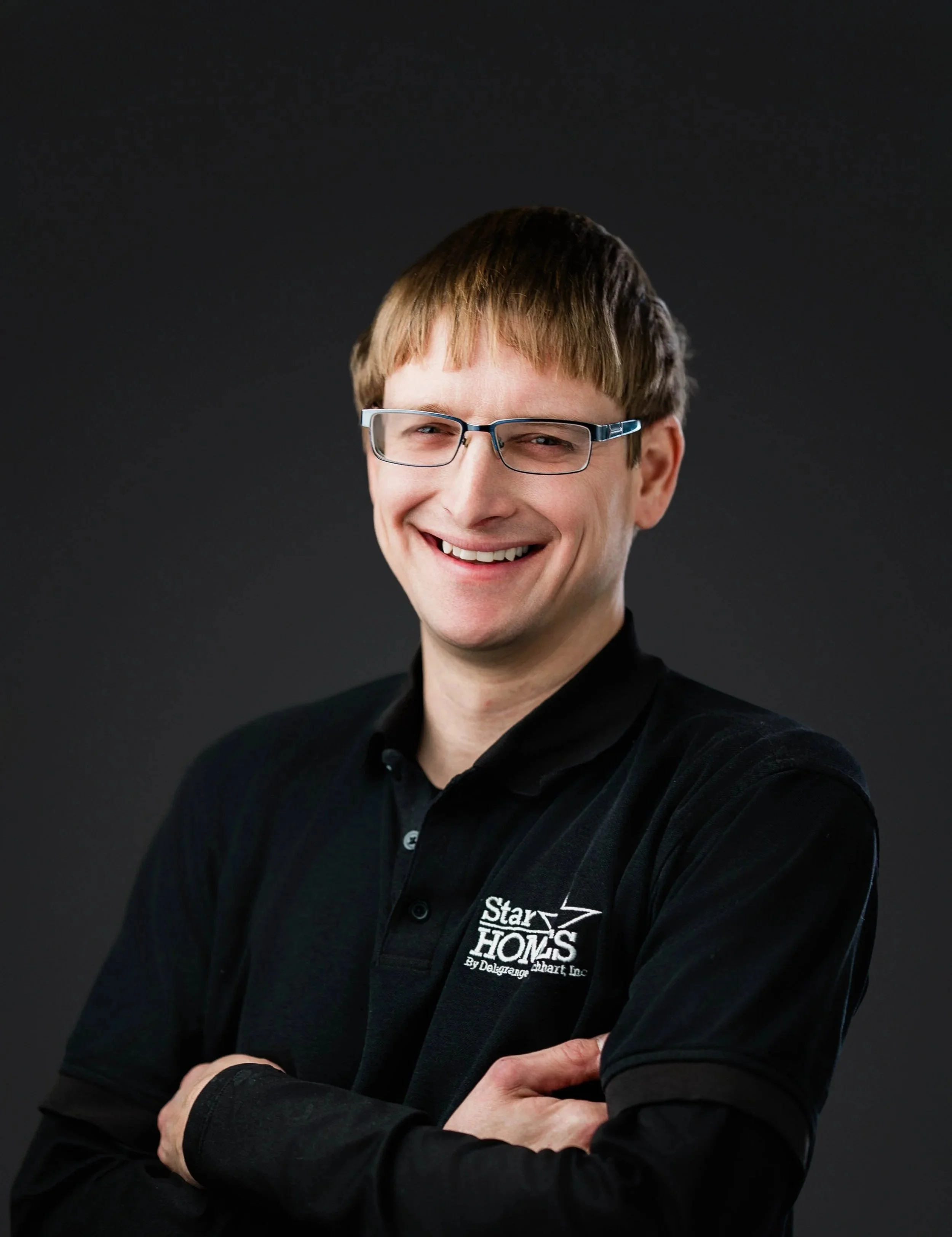 Portrait of a smiling man with light brown hair, wearing glasses and a black shirt with a logo, against a dark background.