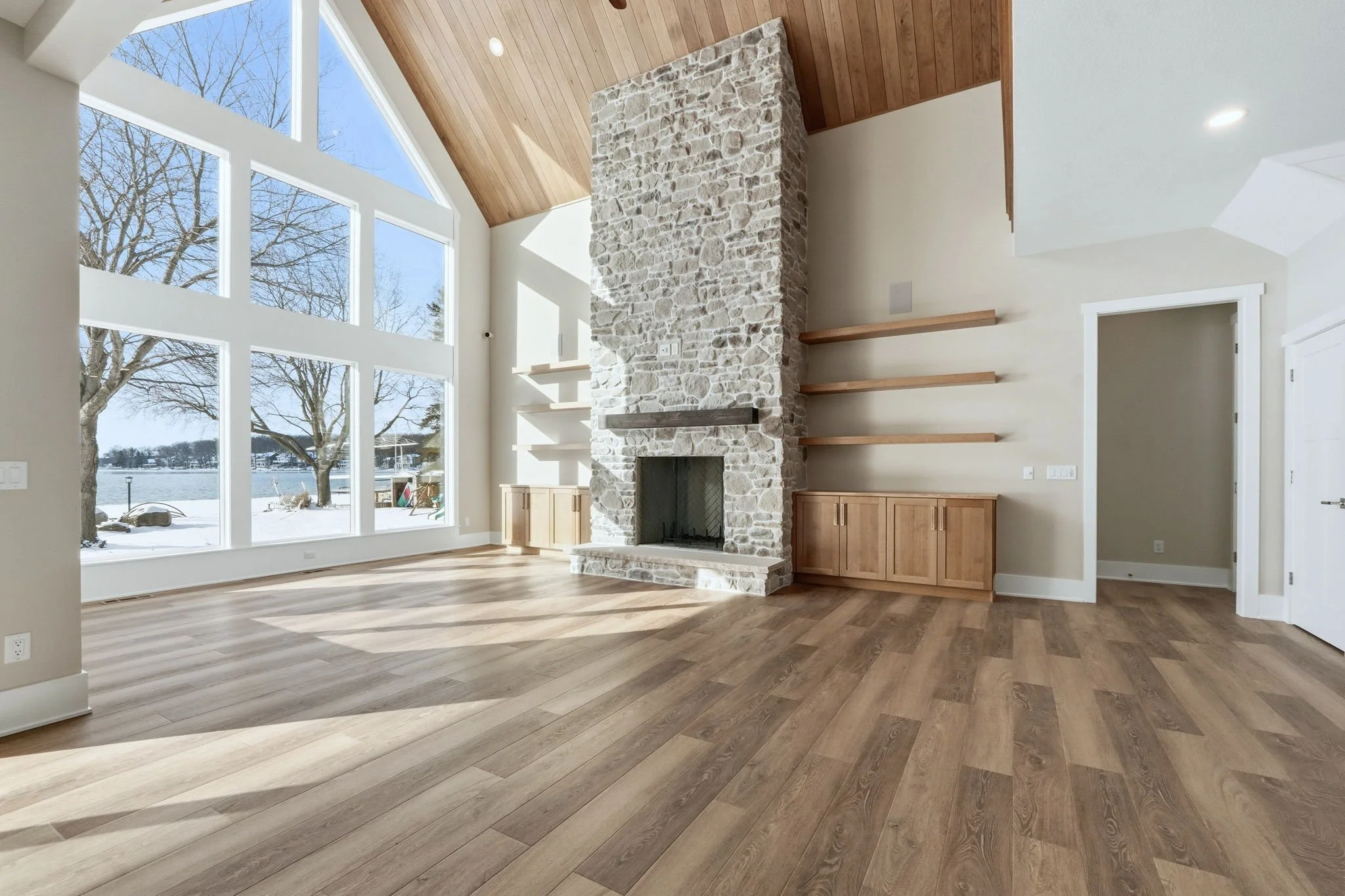 Empty living room with large floor-to-ceiling windows showing trees and water, stone fireplace, built-in wooden cabinetry, and floating shelves, with a wood-paneled vaulted ceiling.