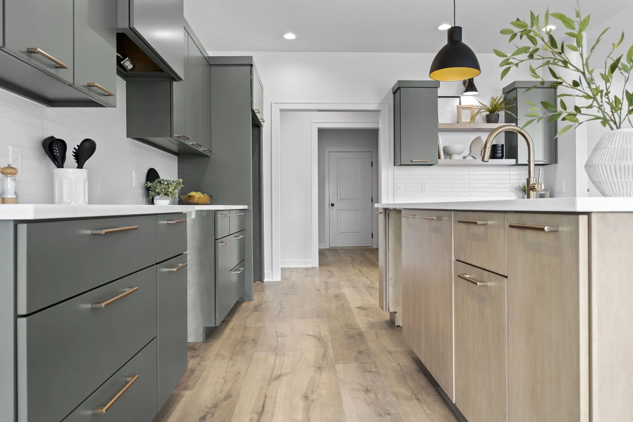 Modern kitchen with green and beige cabinets, white countertops, wood flooring, black hanging light fixture, and decorative shelves with plants and bowls.