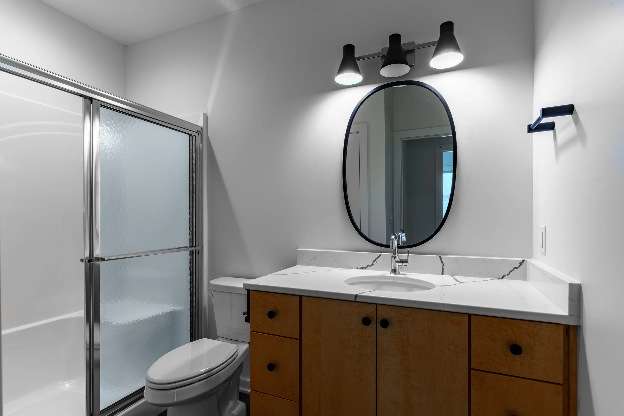 Modern bathroom with white walls, a wooden vanity with a white marble countertop, an oval mirror, black light fixtures, a glass shower door, and a white toilet.