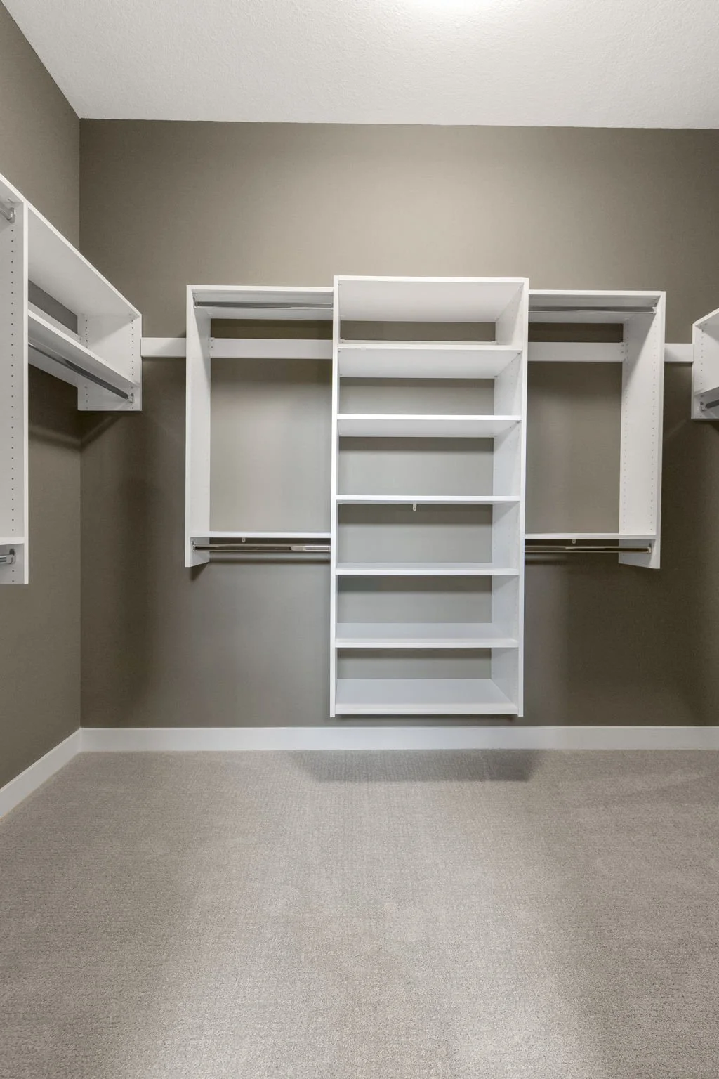 Empty walk-in closet with beige walls, white shelving and hanging rods, and light-colored carpet flooring.