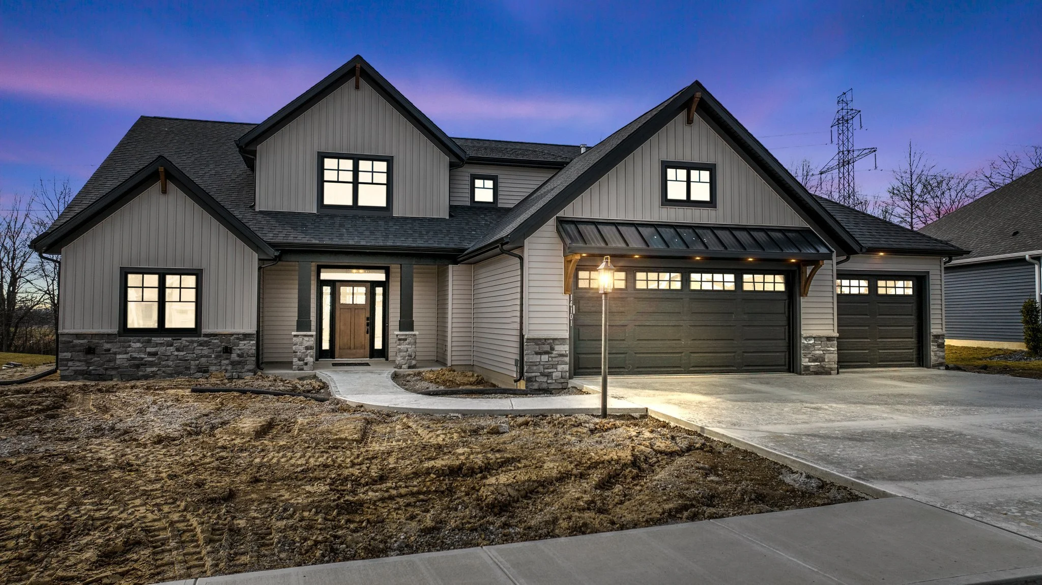 A modern two-story house with gray and white siding, black window frames, and a black garage door, illuminated by a street lamp on a dusk sky background. The front yard is unfinished with dirt and construction materials.