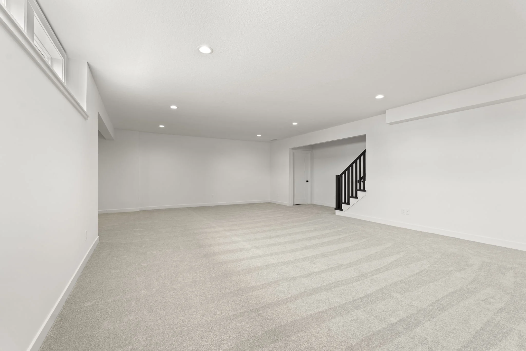 Empty living room with white walls, ceiling lights, light-colored carpet, and a staircase with black railing.