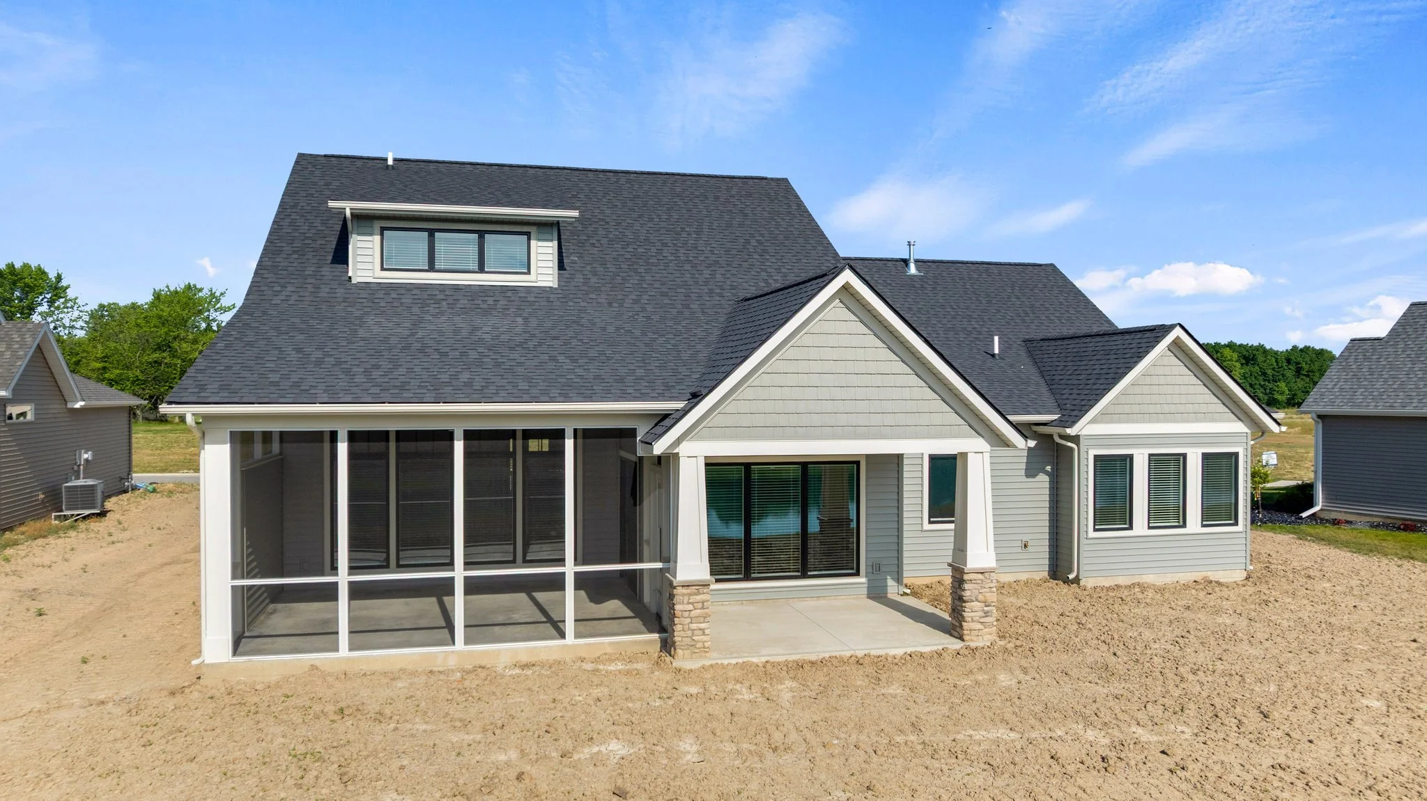 Rear view of a modern house with a screened-in porch, gray siding, and a dark shingle roof, in a newly landscaped yard with neighboring houses and a blue sky in the background.