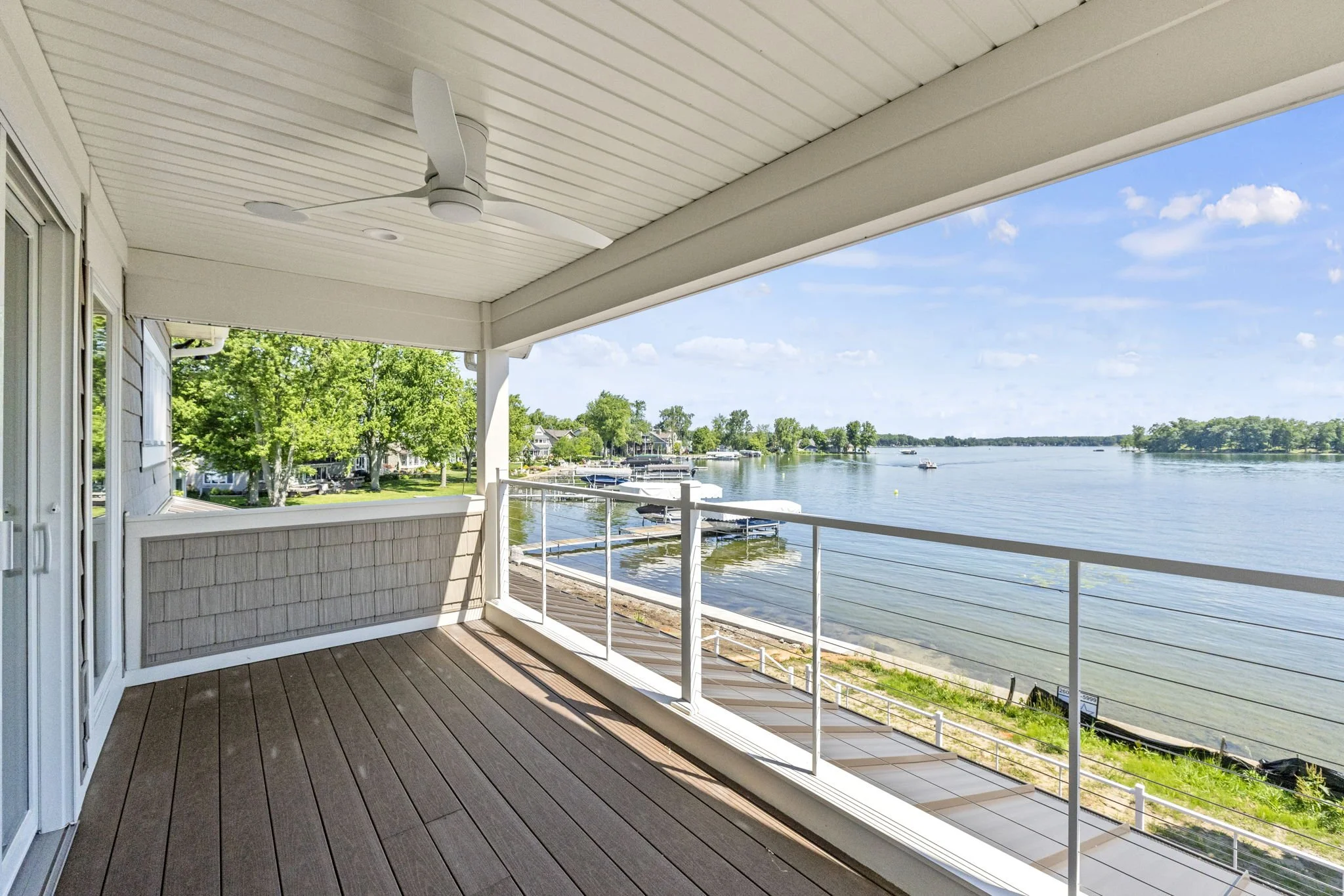 View of a lakeside balcony with wooden flooring, white railing, and a ceiling fan, overlooking a calm lake with boats and trees in the background on a sunny day.