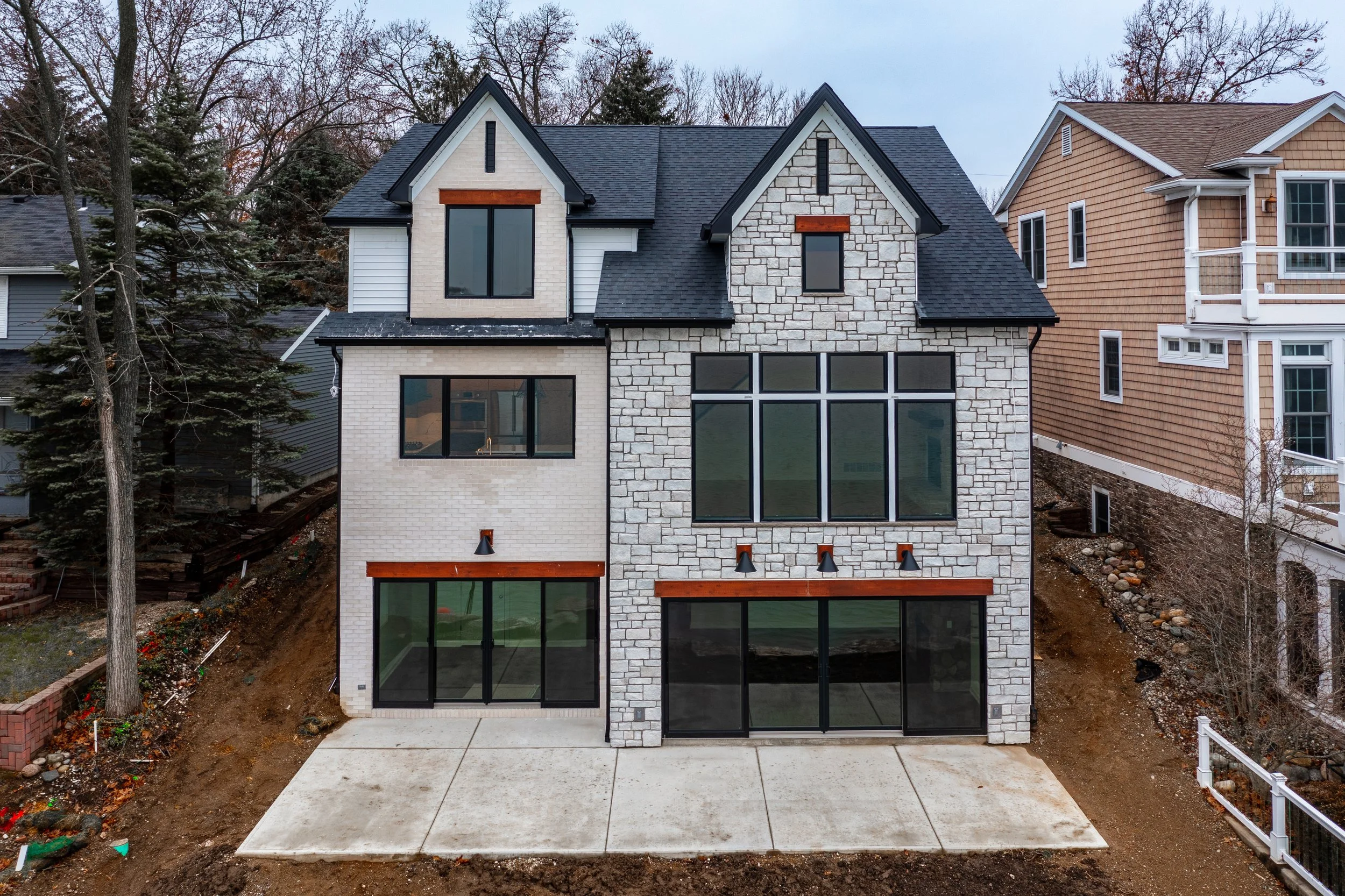 Newly constructed three-story house with a stone and white brick exterior, large black-framed windows, a black shingle roof, and a concrete driveway in front, neighboring houses on either side, and trees in the background.