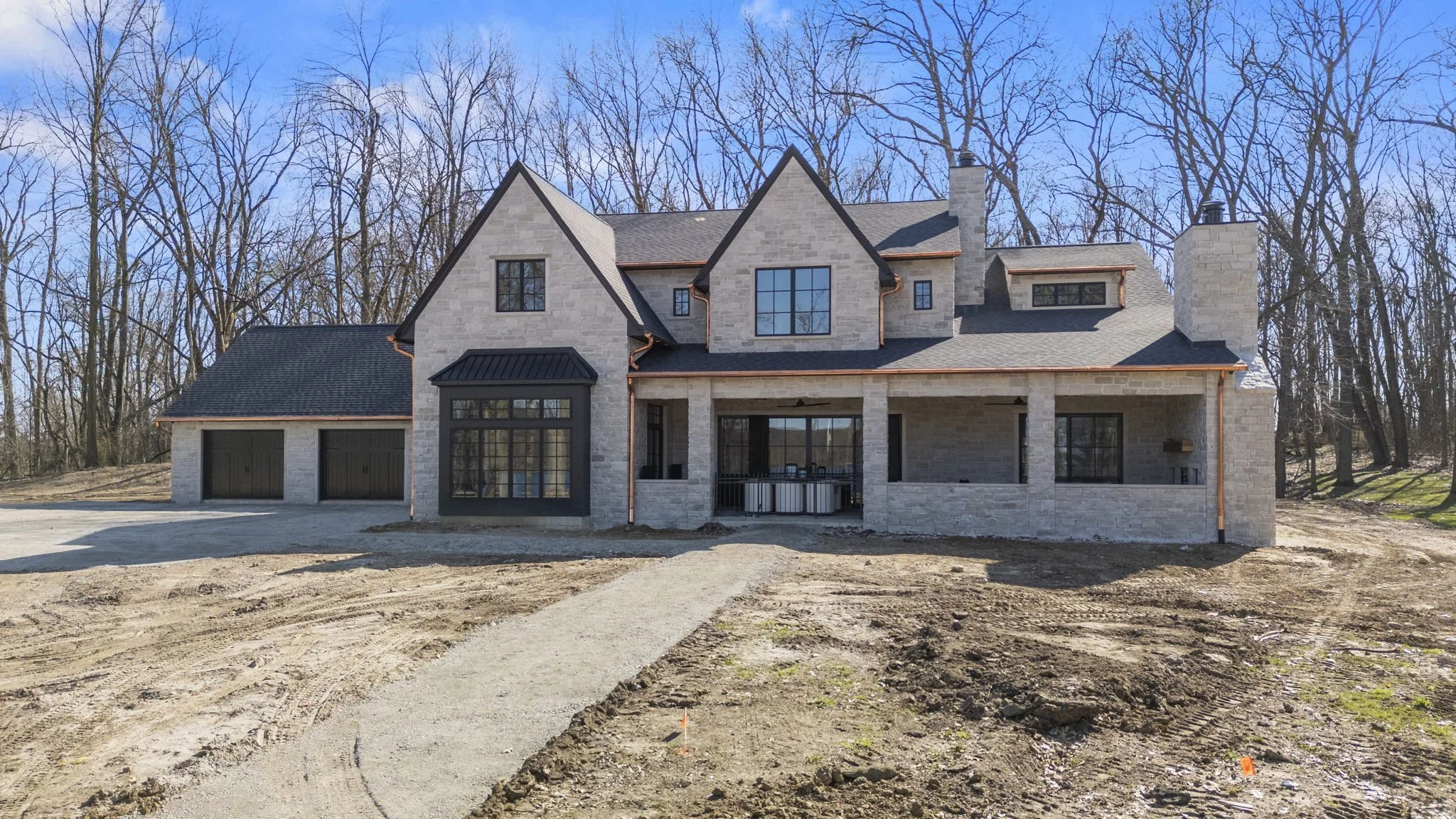 Newly constructed house with stone exterior, black trim, large front porch, and a spacious driveway in a wooded area.