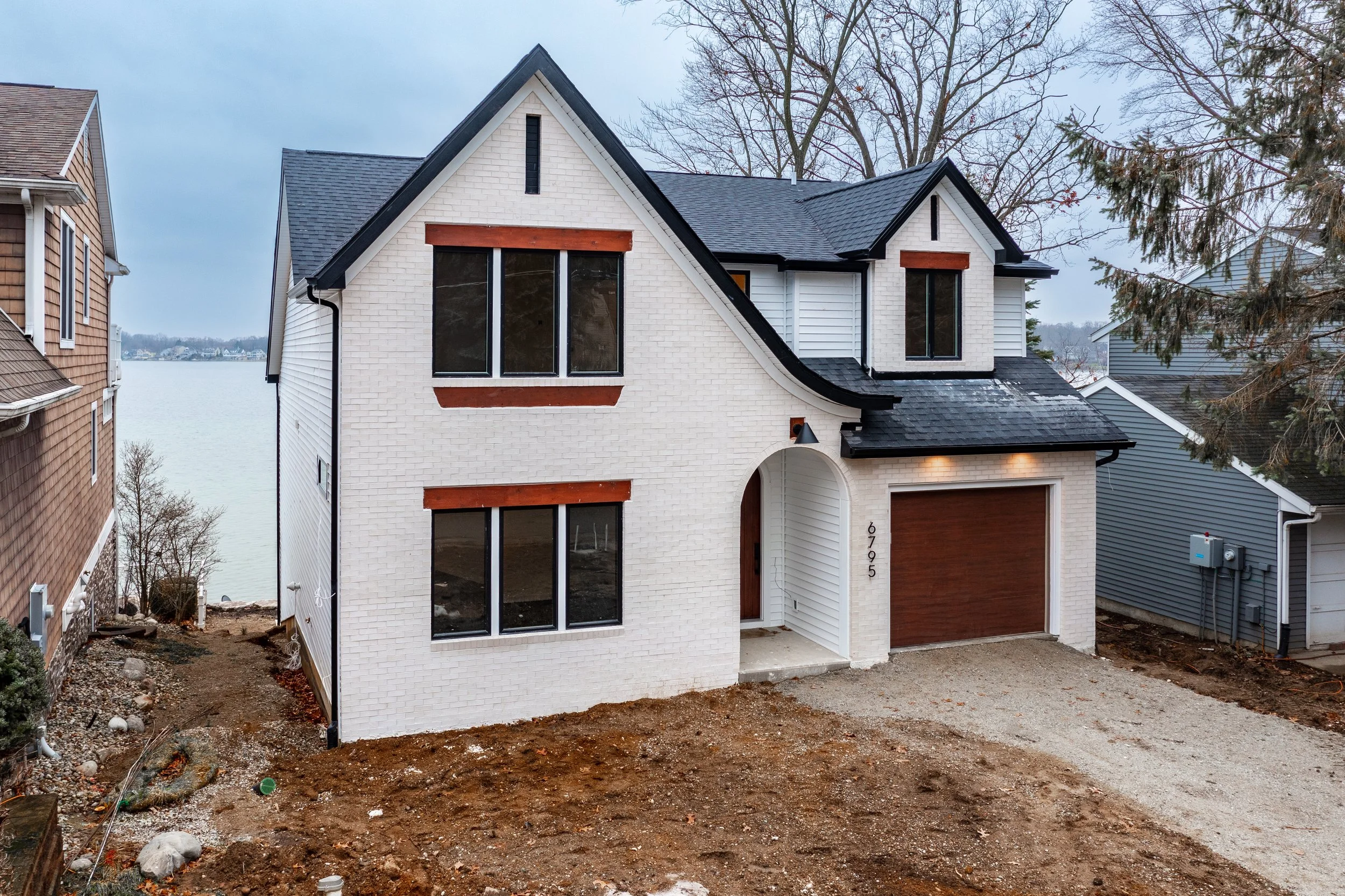 New white two-story house with black roof, brown window frames, and a brown garage door, situated on a dirt lot near a body of water, with neighboring houses on either side.