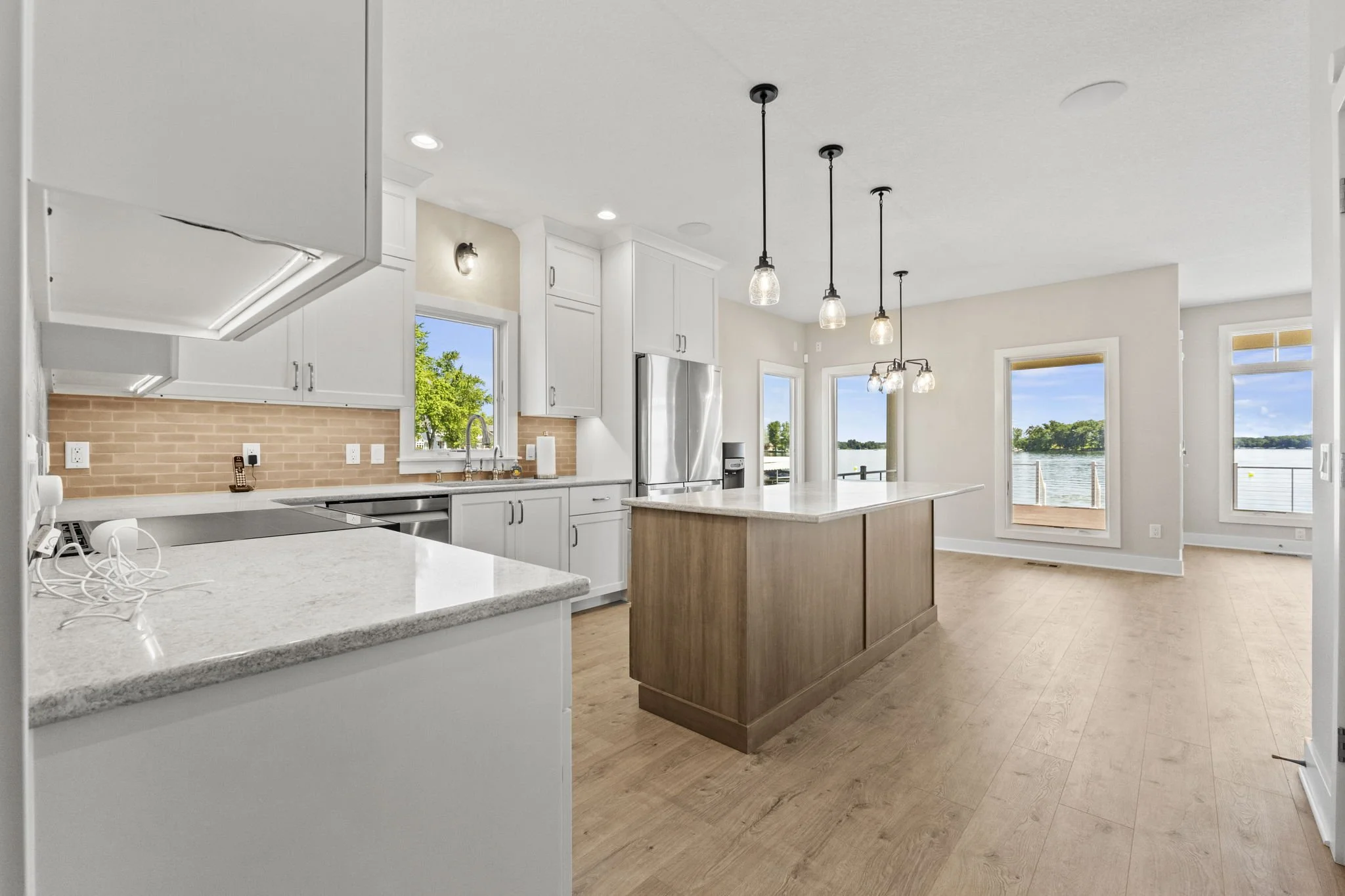 Bright, modern kitchen with white cabinets, a large island with a marble countertop, pendant lights, stainless steel refrigerator, and large windows overlooking a lake.