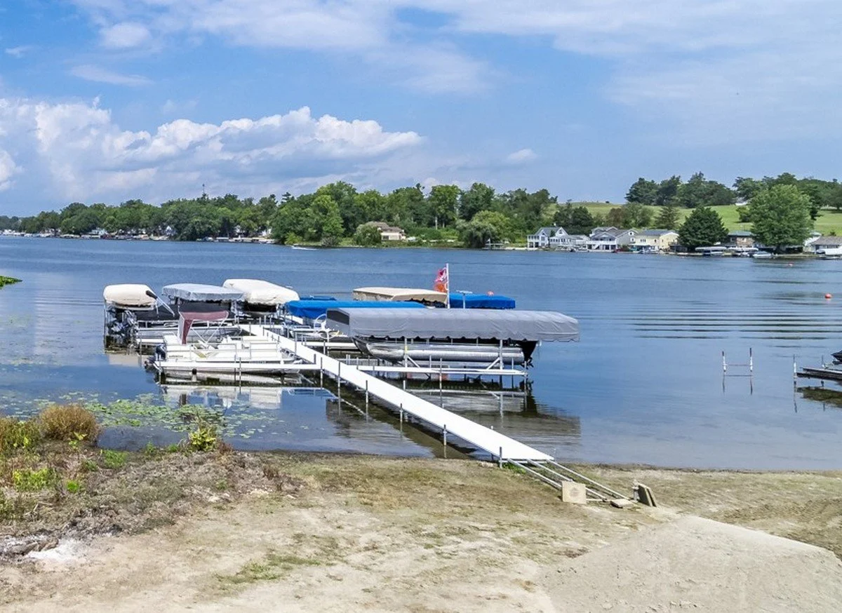 Dock with covered boat slips on a lake, surrounded by houses and trees under a partly cloudy sky.