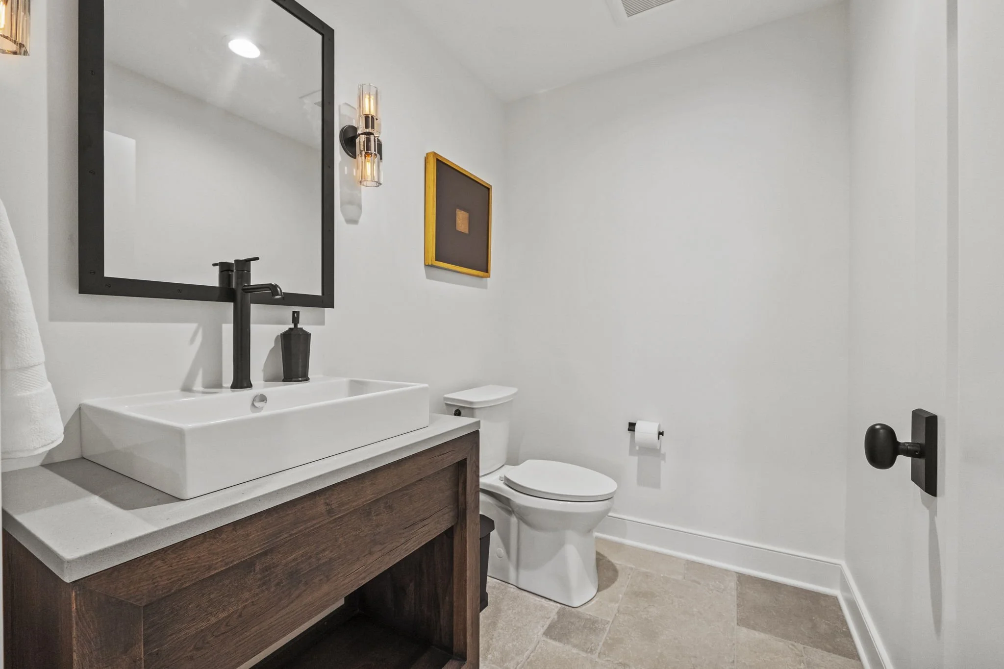 Modern bathroom with white walls, a wooden vanity with a white vessel sink, a black faucet, a large mirror, a black soap dispenser, a wall-mounted light fixture, a framed artwork, a white toilet, and a toilet paper roll.