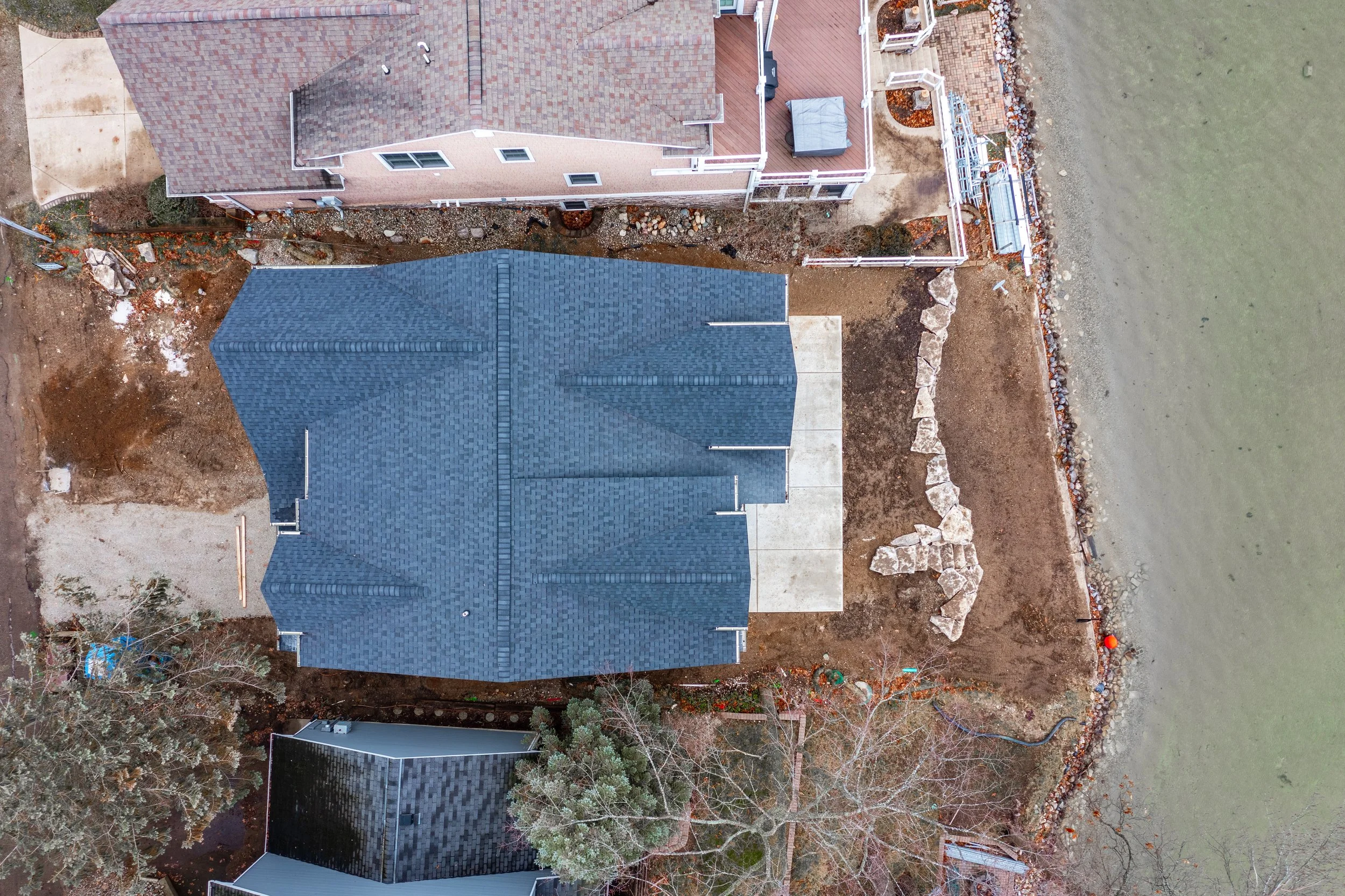 Aerial view of a house with a blue roof next to a body of water, with neighboring houses, trees, and a rocky shoreline.