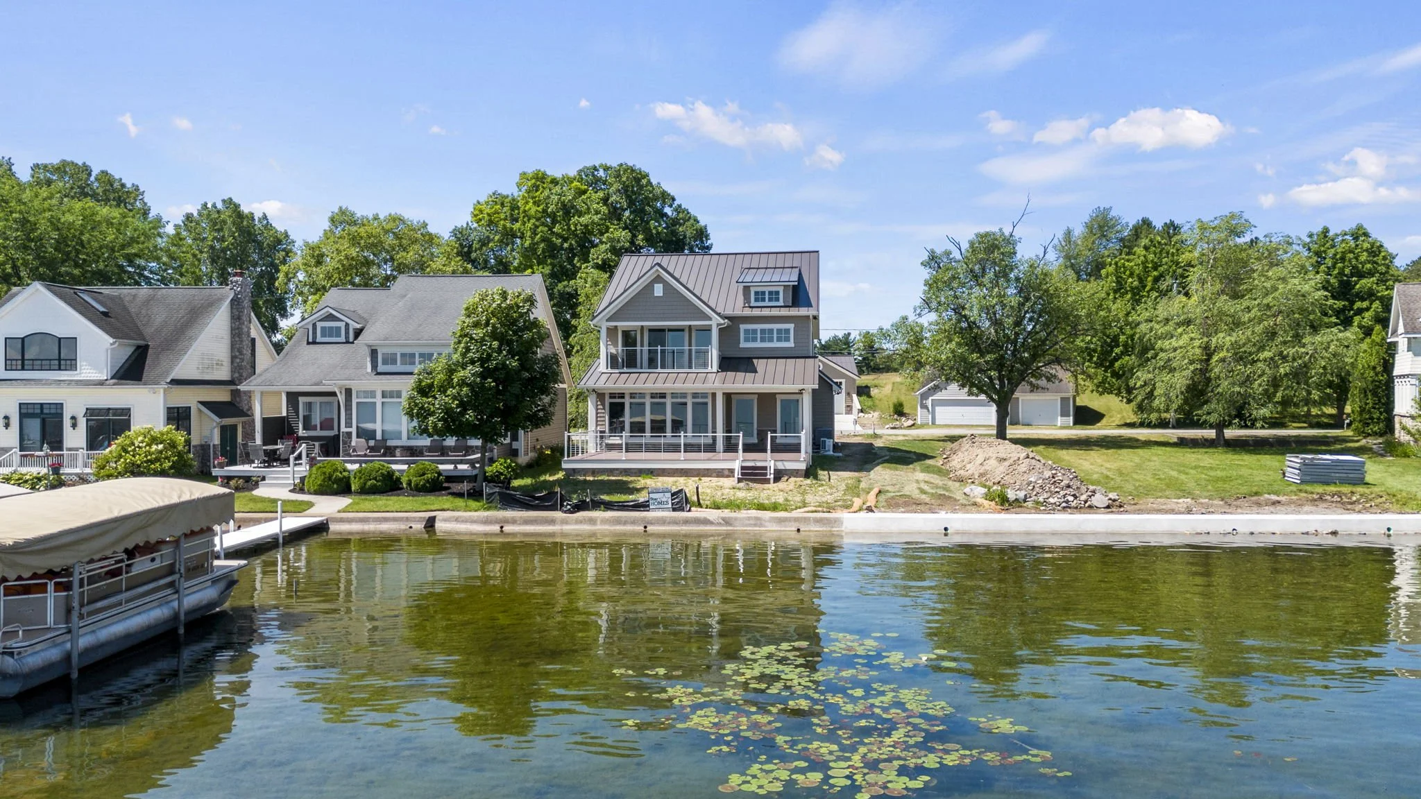 Waterfront view of houses and trees along a calm lake under a blue sky with white clouds.