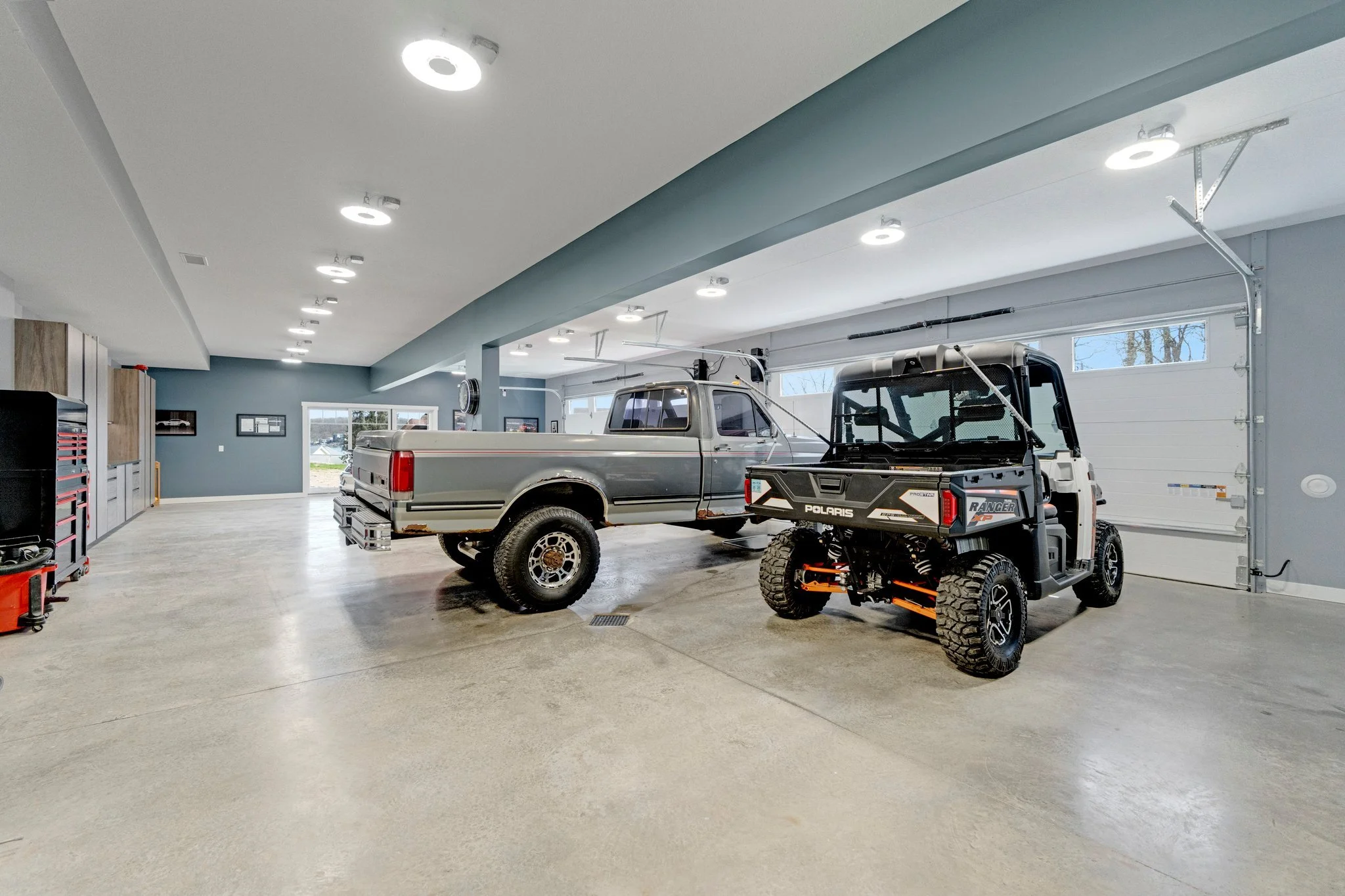 A spacious garage with a gray pickup truck and a black Polaris off-road vehicle parked inside. The garage has gray walls, large windows, and overhead lighting.