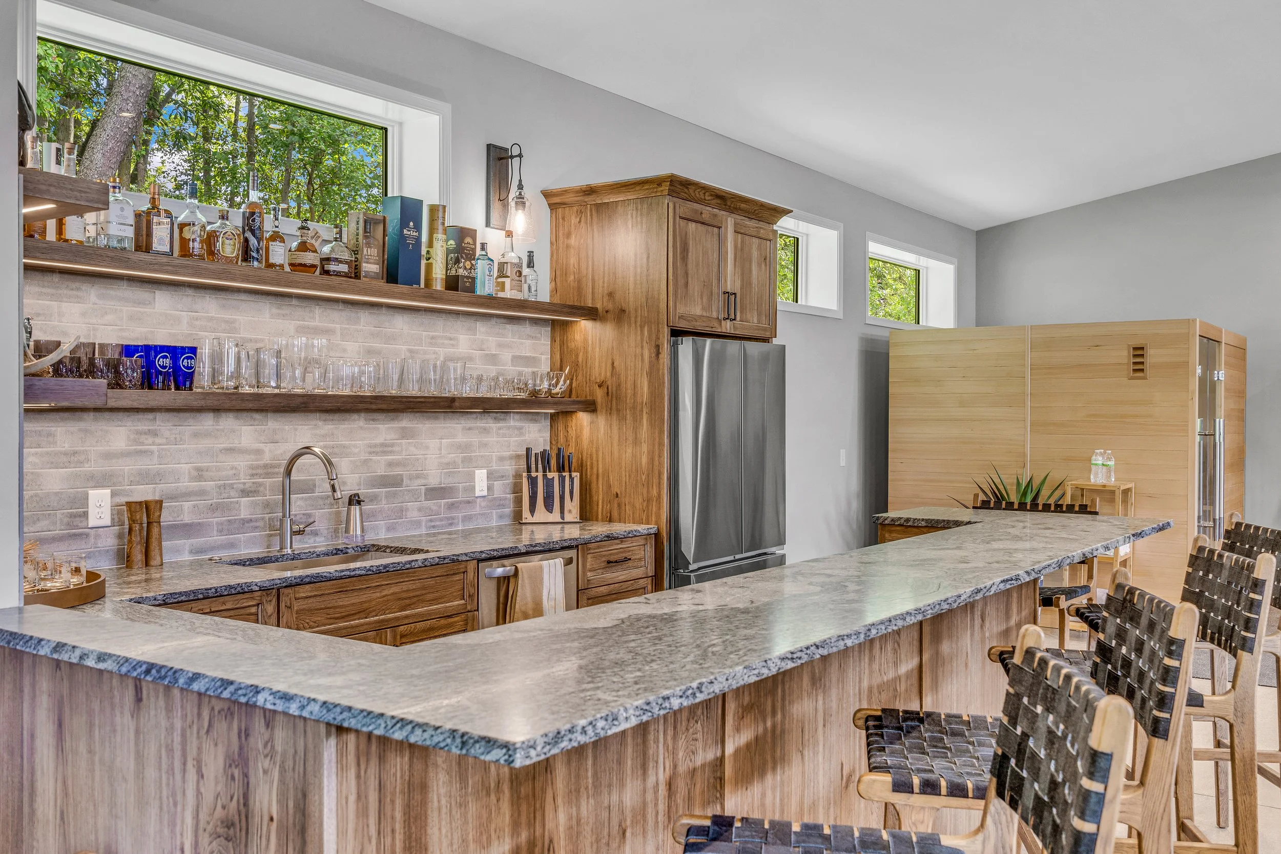Modern kitchen with granite countertop bar, wooden cabinetry, open shelves with glassware and liquor bottles, stainless steel refrigerator, and woven barstools.