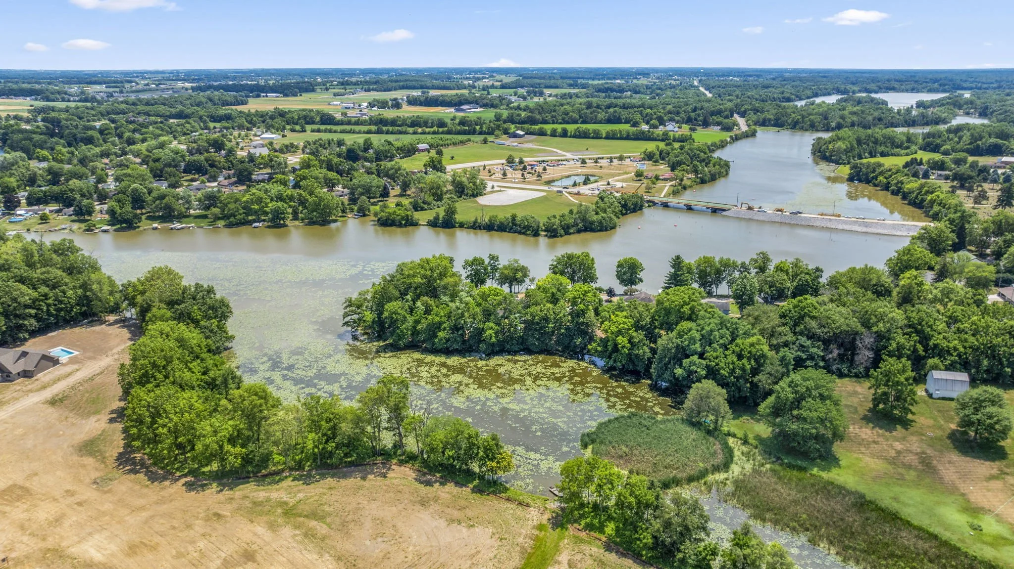 Aerial view of a winding river surrounded by lush green trees, residential areas, and open land with a bridge across the river under a partly cloudy sky.