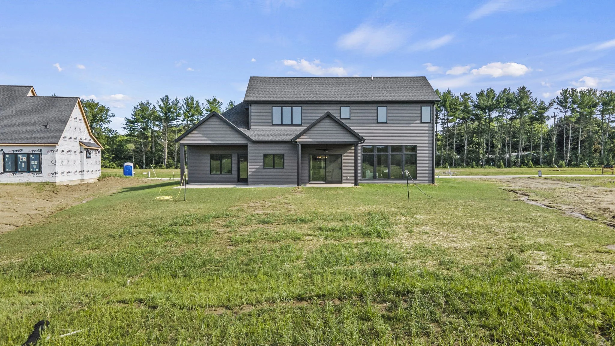 Newly built gray house with large windows on a grassy lot under a blue sky with some clouds, neighboring houses under construction on the left, and trees in the background.