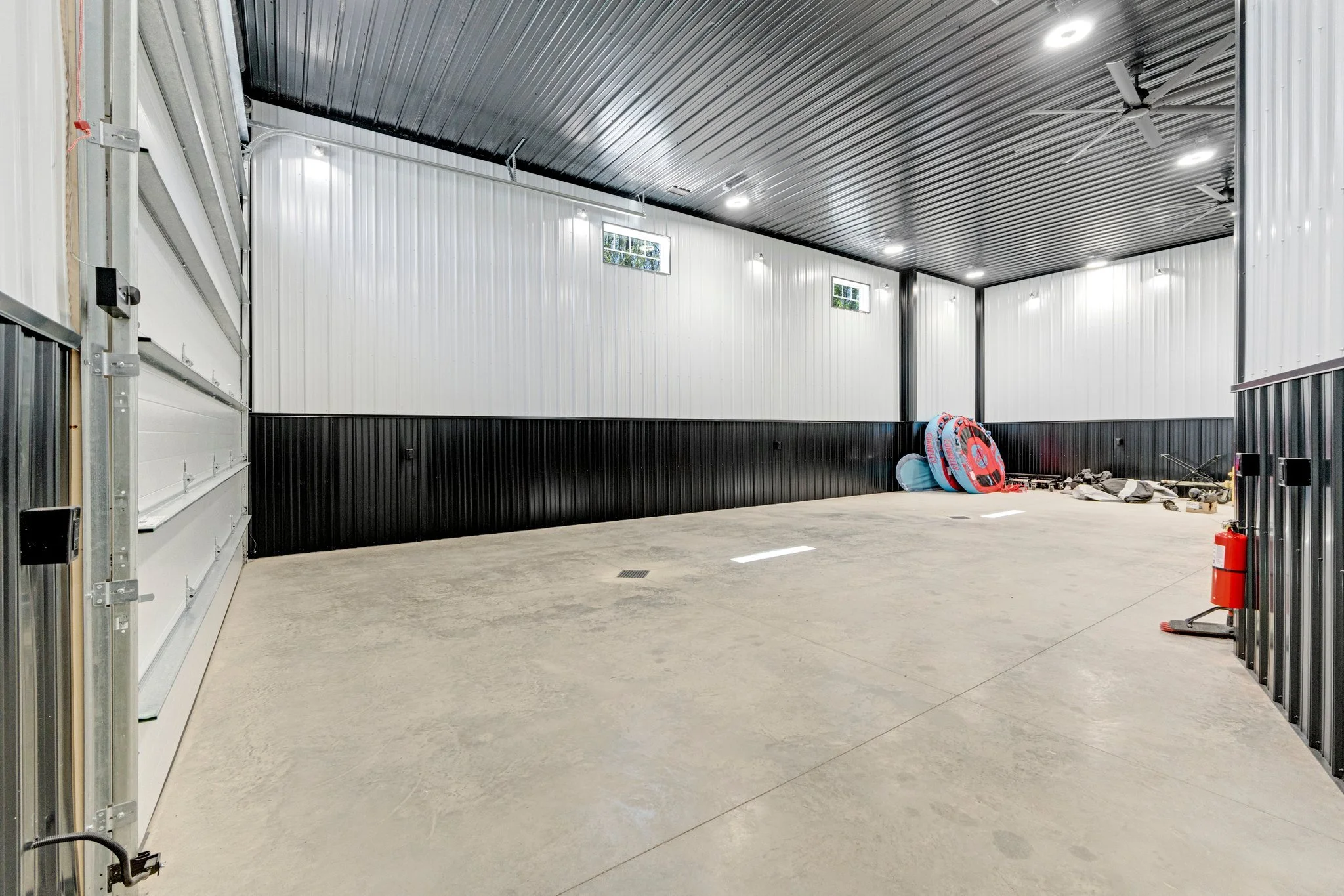 Empty garage with concrete floor, white and black metal walls, a dark metal ceiling with ceiling fans, small windows near the ceiling, and some inflatable tubes and rocks in the corner.