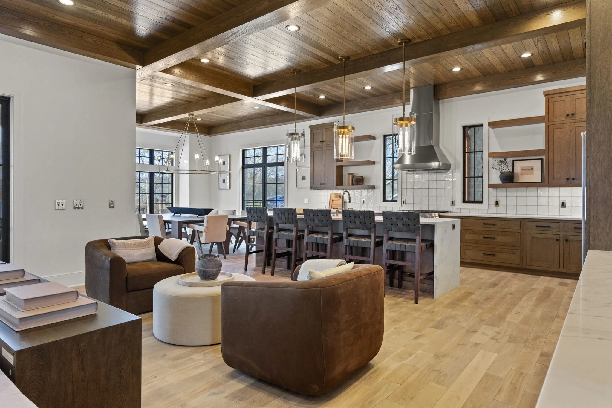 Open-concept living space with a living room and kitchen featuring wooden ceiling beams, pendant lighting, brown leather armchairs, a white ottoman, a wooden dining table with beige chairs, and a kitchen island with six dark wood barstools.