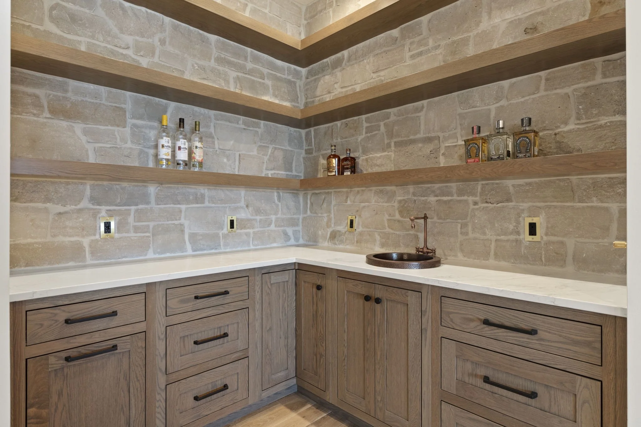 Kitchenette with beige wooden cabinets, a copper sink, and brick walls with floating wooden shelves holding liquor bottles.